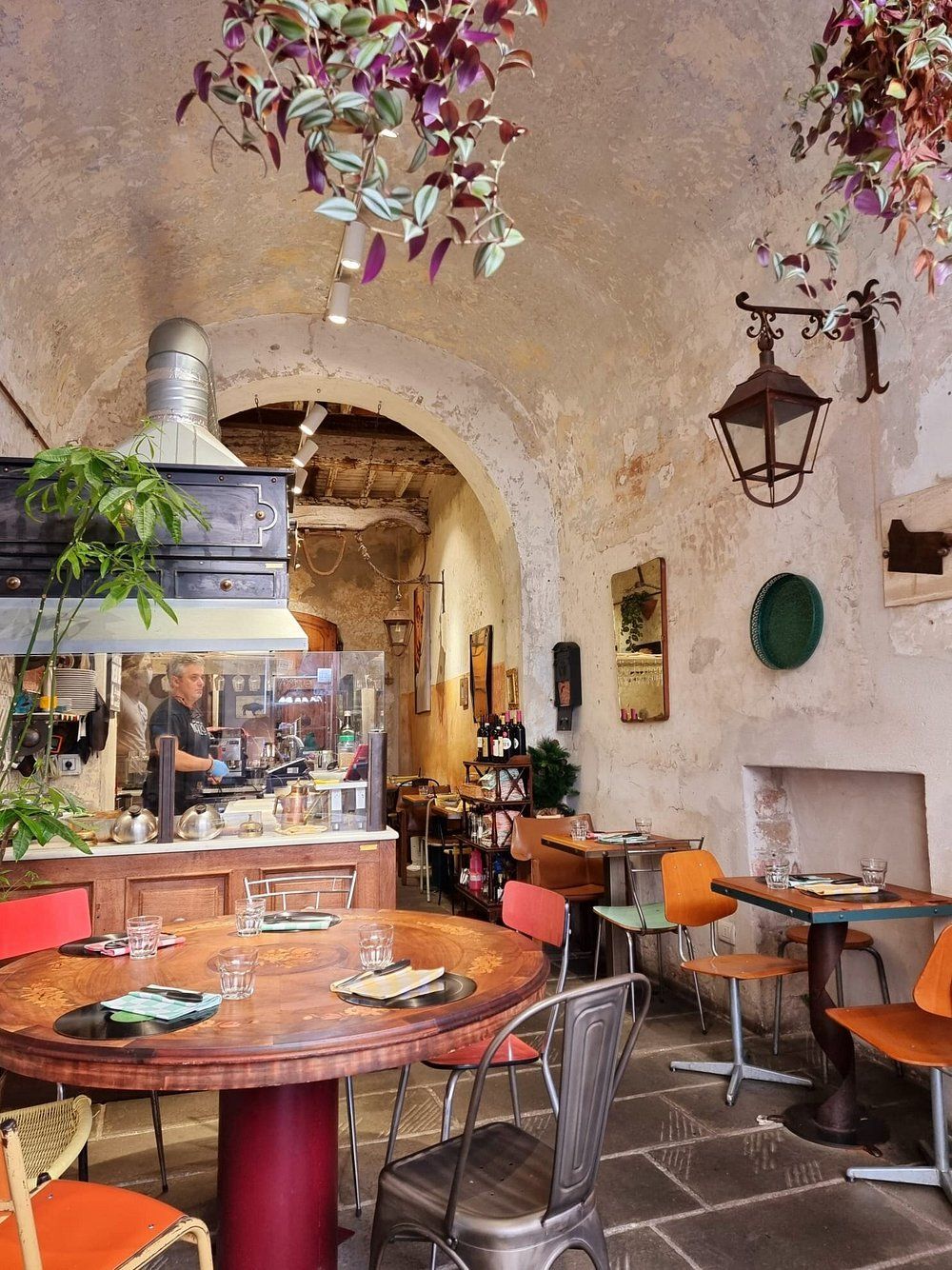 Accogliente ristorante interno con tavoli rotondi, sedie colorate e uno chef al lavoro nella cucina a vista. Soffitto a volta con piante sospese.