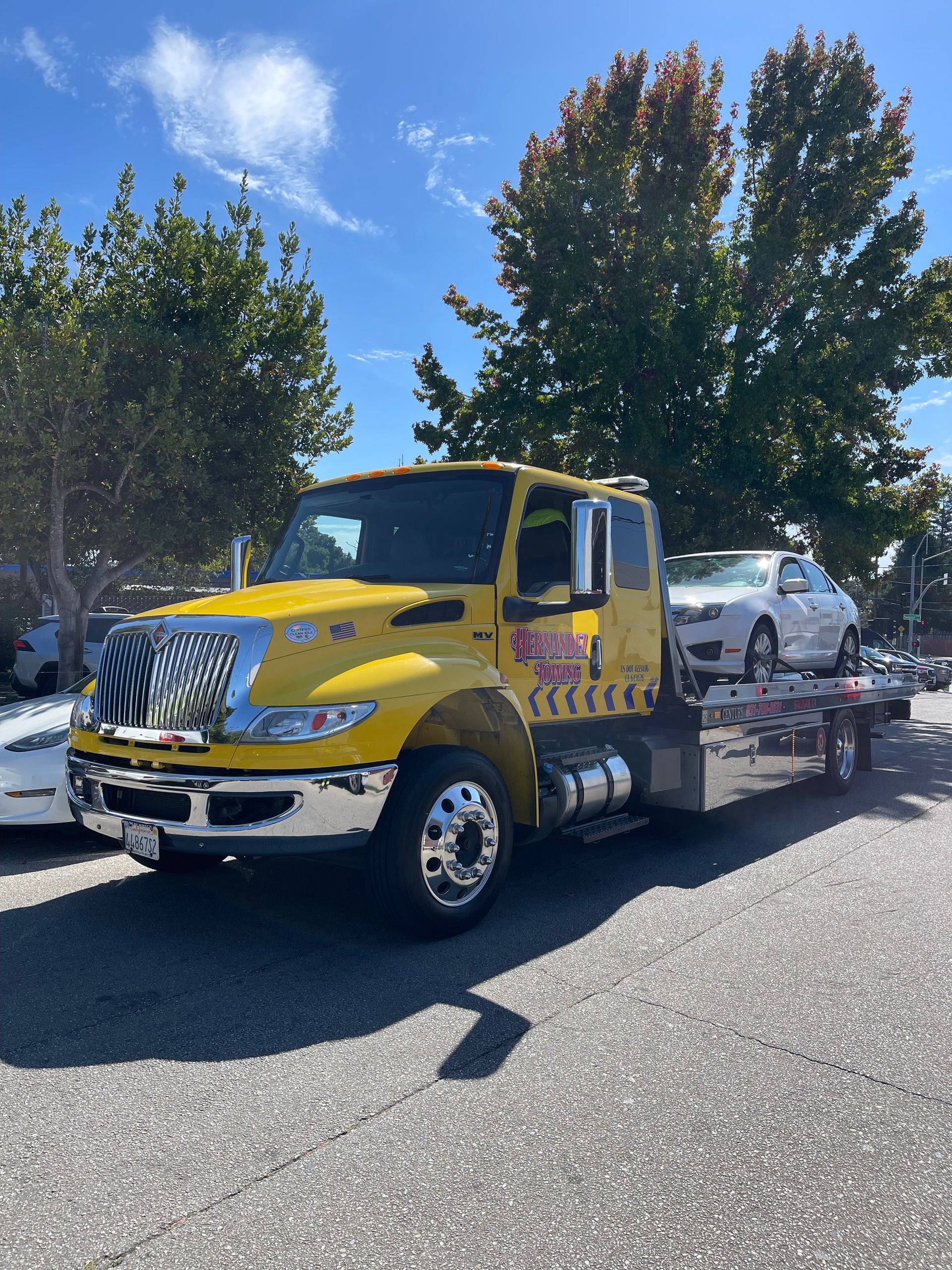 A green car being towed by a tow truck; several people and other cars are present on a paved area.