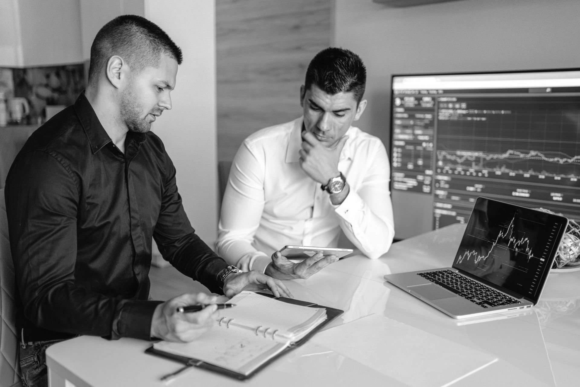 Two men are sitting at a table with a laptop and a clipboard.