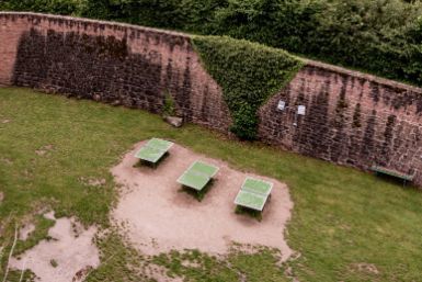 Drei grüne Tischtennisplatten auf einem unbefestigten Fleckchen Erde in einer Rasenfläche, umgeben von einer Backsteinmauer und Vegetation.