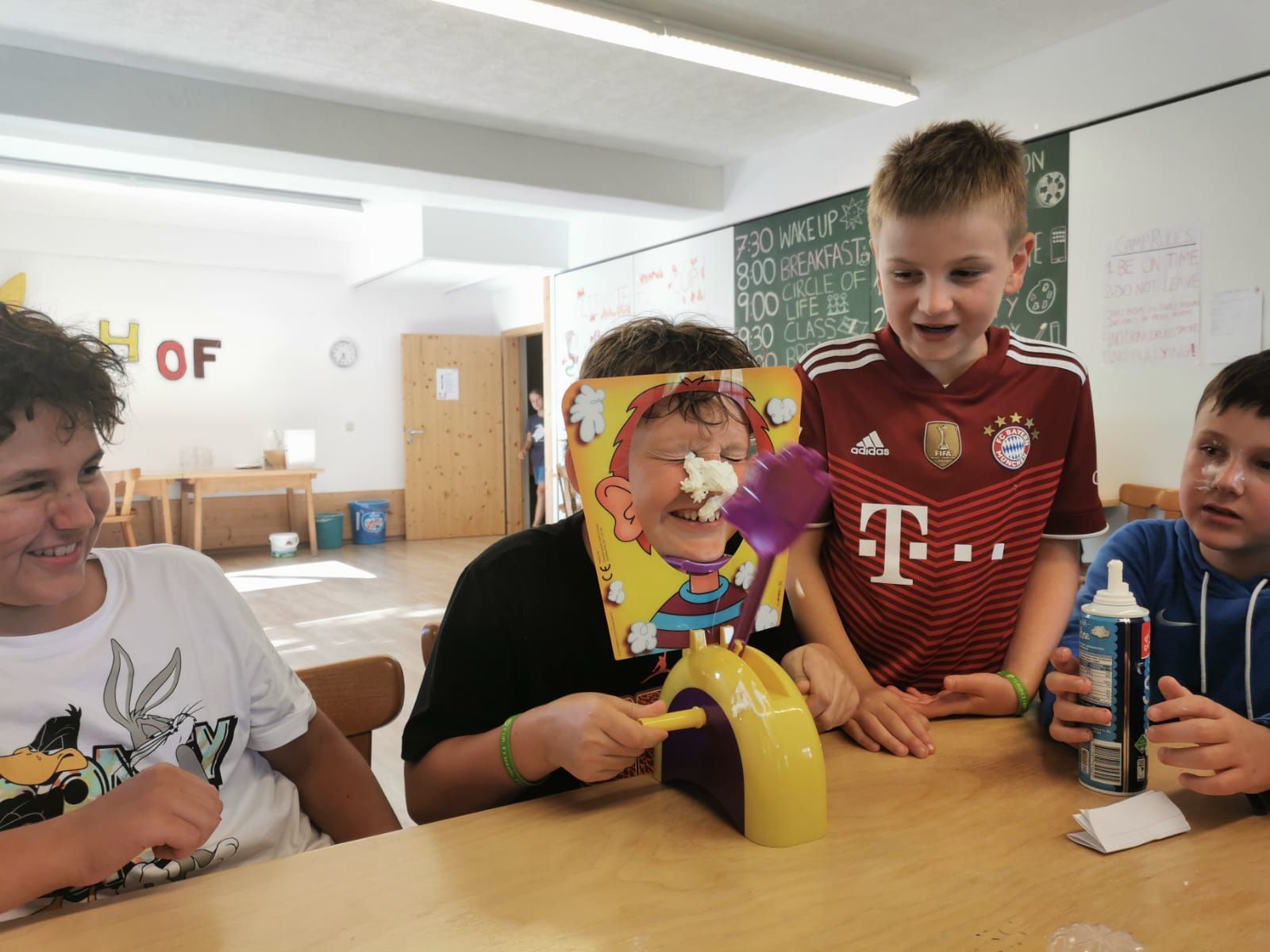 Vier Kinder spielen ein „Kuchengesicht“-Spiel. Eines hat eine Gesichtskarte mit Sahne im Gesicht. Die anderen schauen zu, lächeln und lachen.