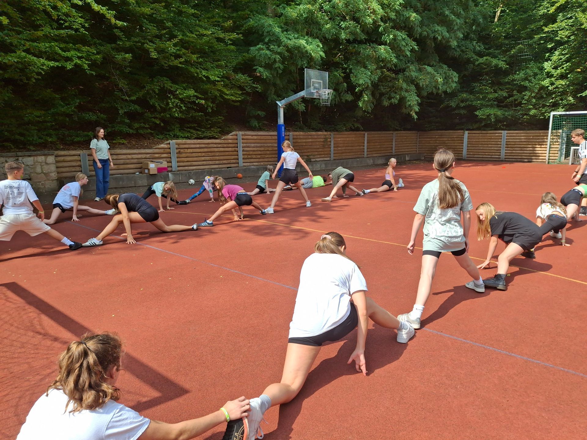 Eine Gruppe Kinder dehnt sich auf einem roten Freiplatz in der Nähe eines Basketballkorbs. Sonnenlicht strömt herein.