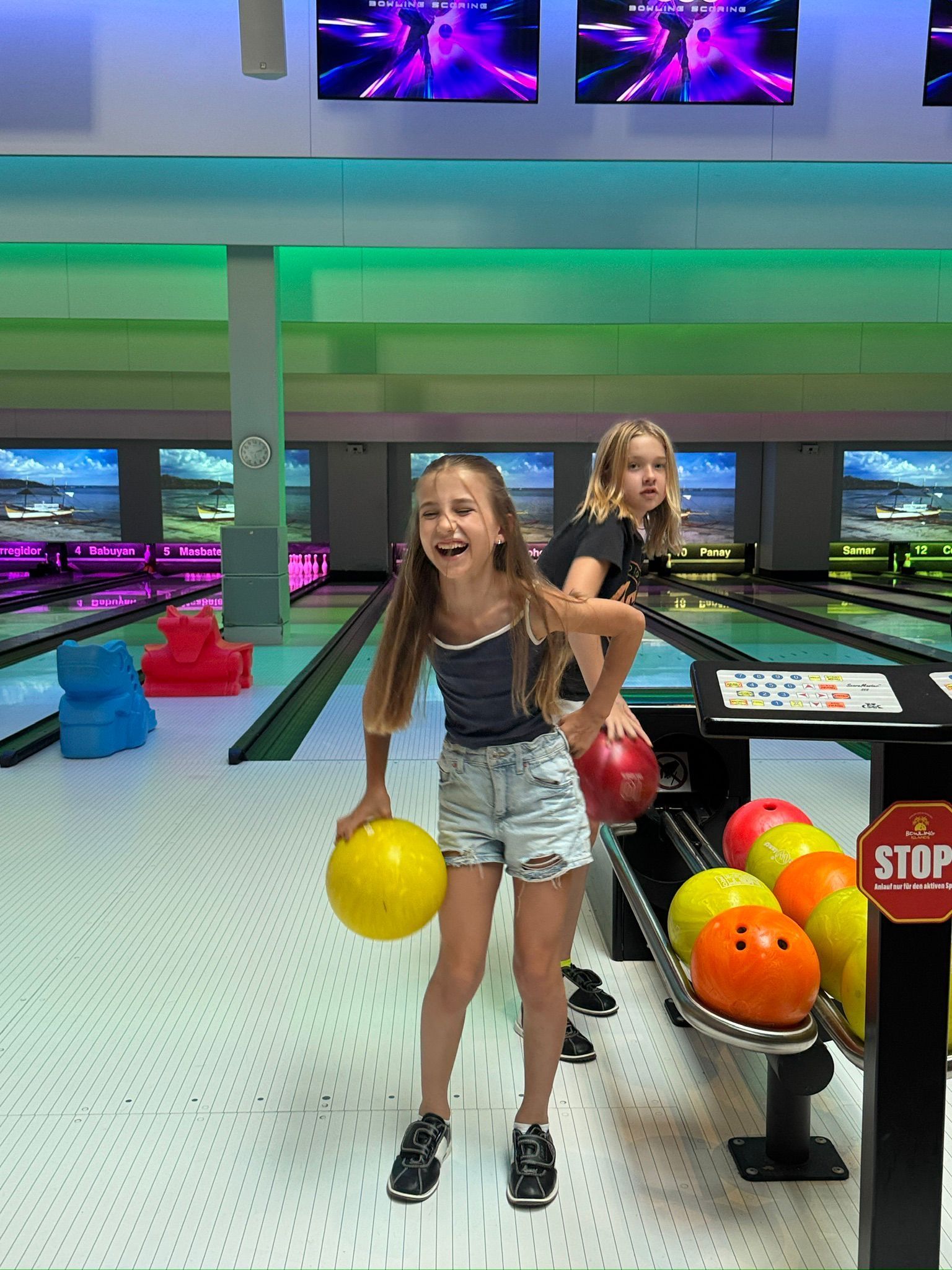 Zwei Mädchen in einer Bowlingbahn, eines hält eine gelbe Kugel und lächelt. Grüne und violette Lichter, im Hintergrund die Bowlingbahnen.