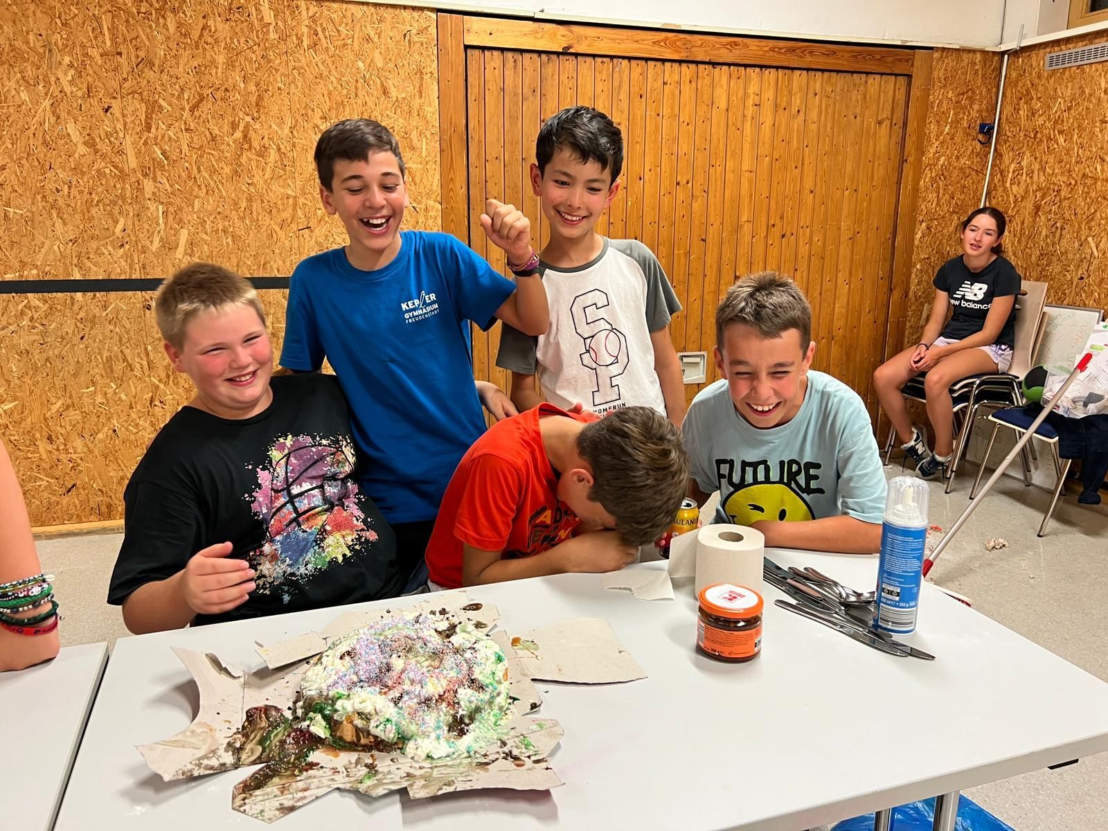 Eine Gruppe Jungen sitzt an einem Tisch mit einem Kuchen. Einige lachen, einer hat sein Gesicht im Kuchen.