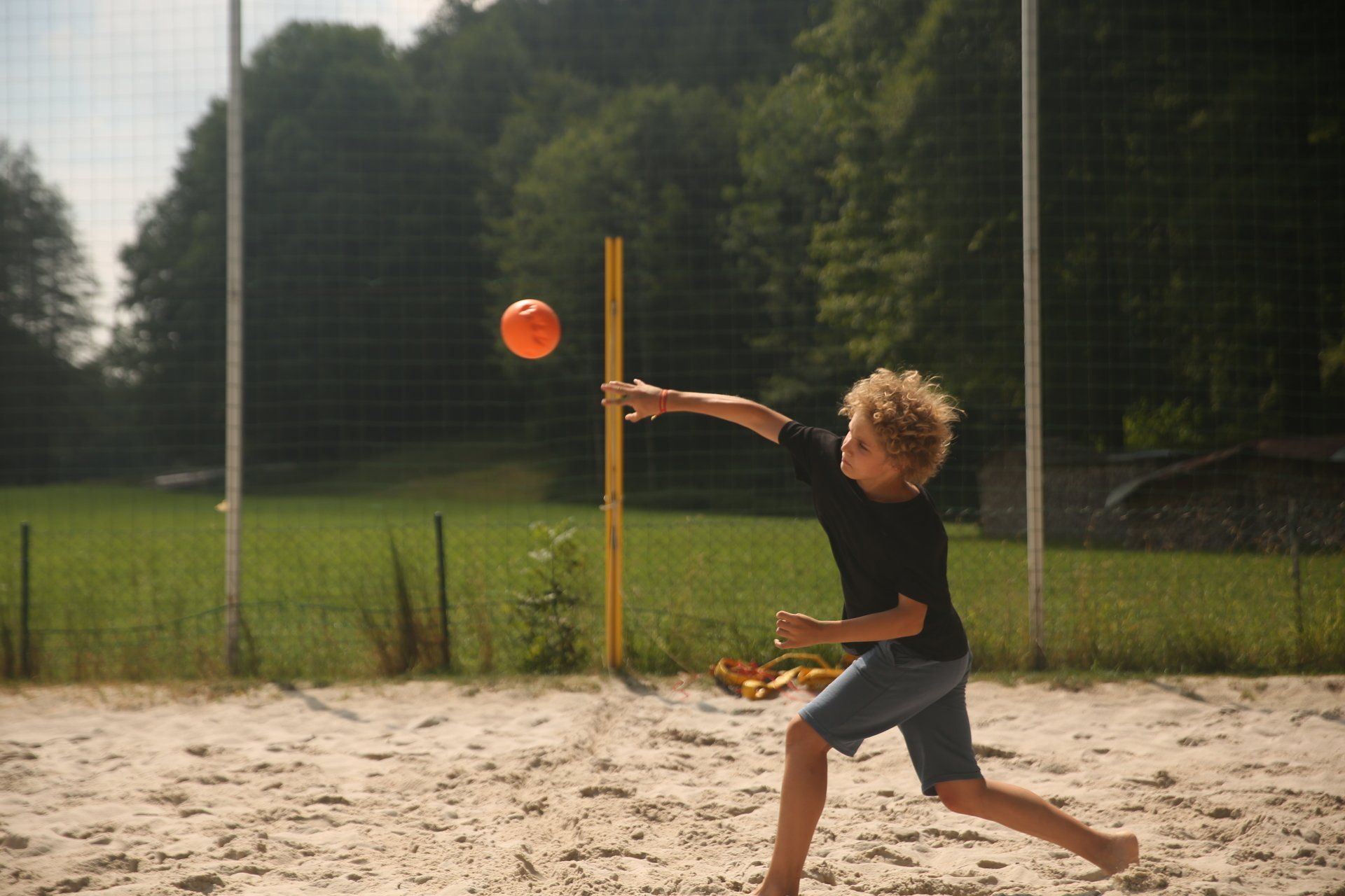 Junge wirft einen orangefarbenen Ball auf einen Sandplatz; im Hintergrund sind Bäume zu sehen.