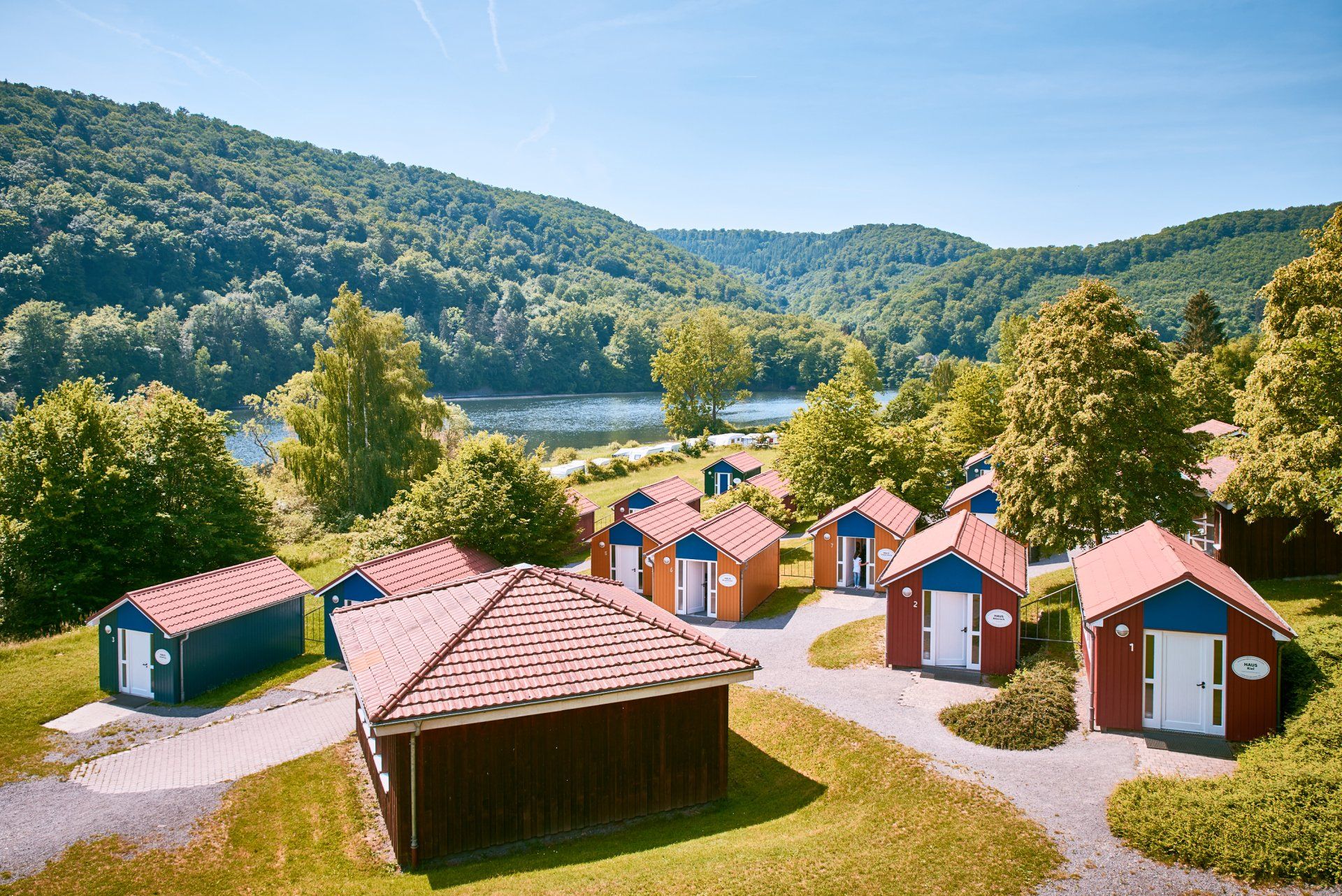Hütten mit roten Ziegeldächern in der Nähe eines Sees und bewaldete Hügel unter blauem Himmel.