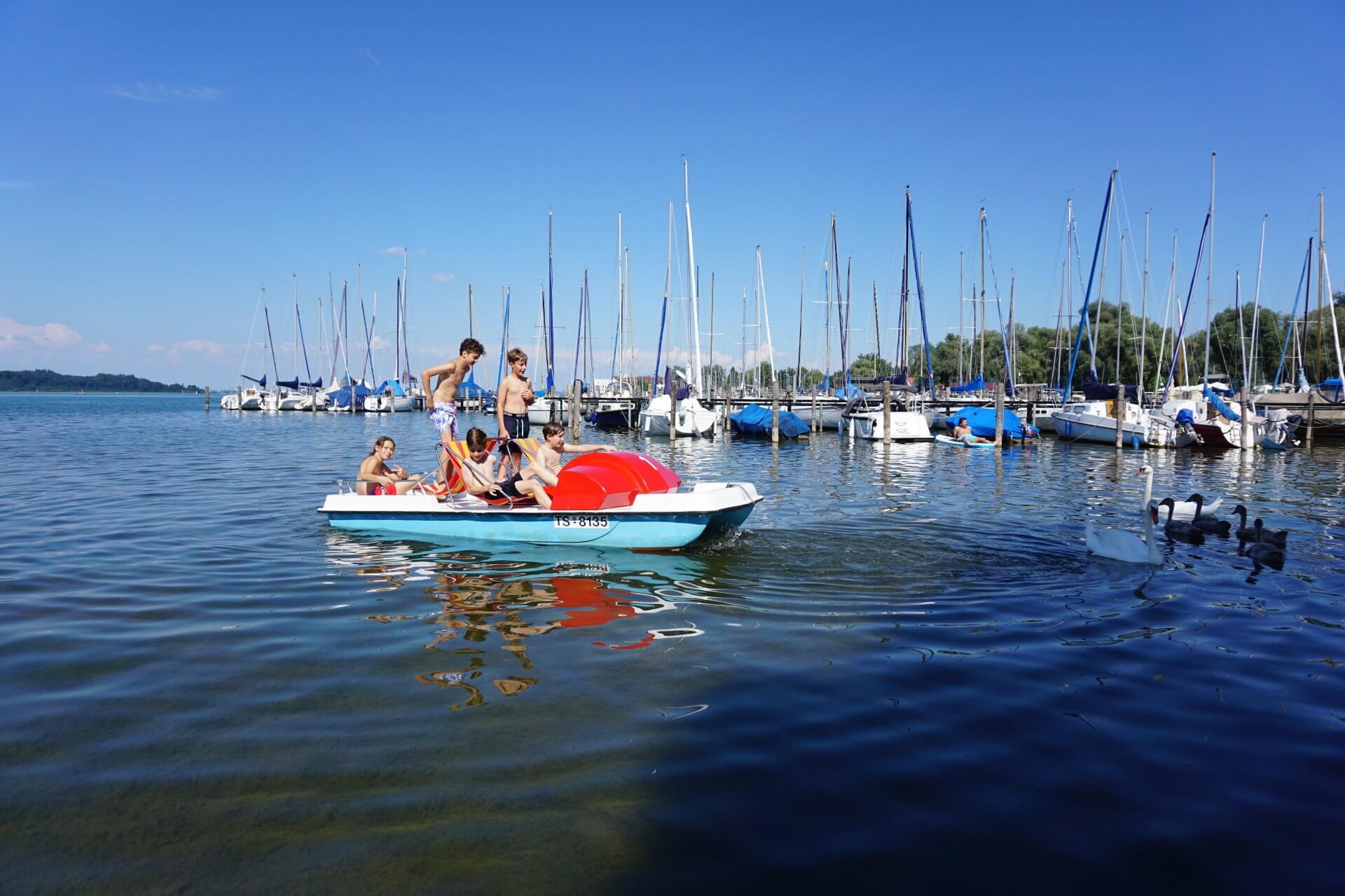 Menschen auf einem blauen Paddelboot in einem Hafen, im Hintergrund Segelboote unter blauem Himmel.