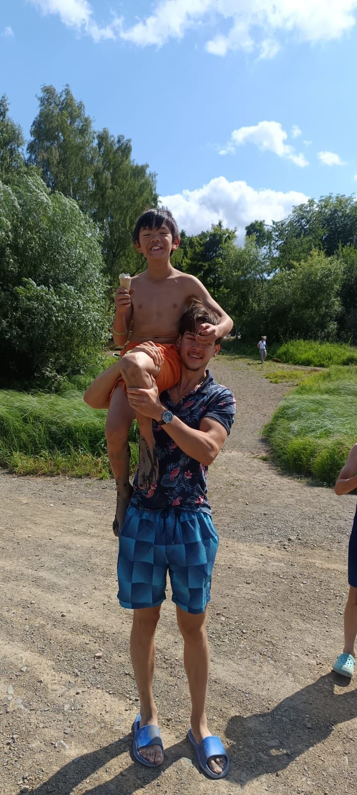 An einem sonnigen Tag trägt ein Mann einen Jungen auf seinen Schultern. Sie stehen auf einem Feldweg in der Nähe von Bäumen.