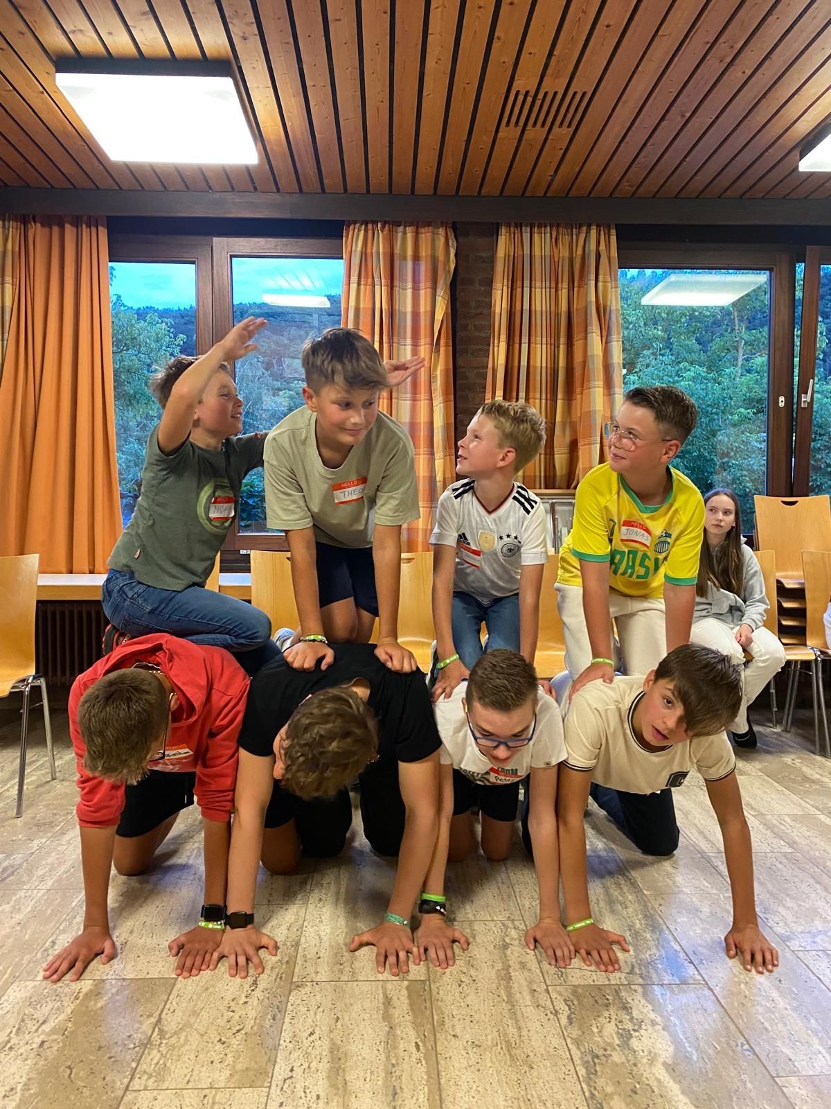 Kinder bauen drinnen eine Menschenpyramide, lächeln und tragen verschiedenfarbige Hemden.