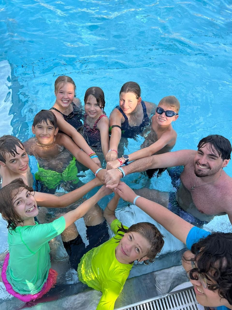 Eine Gruppe von Menschen in einem Pool, die Hände im Kreis. Lächelnde Gesichter, blaues Wasser, sonnige Umgebung.