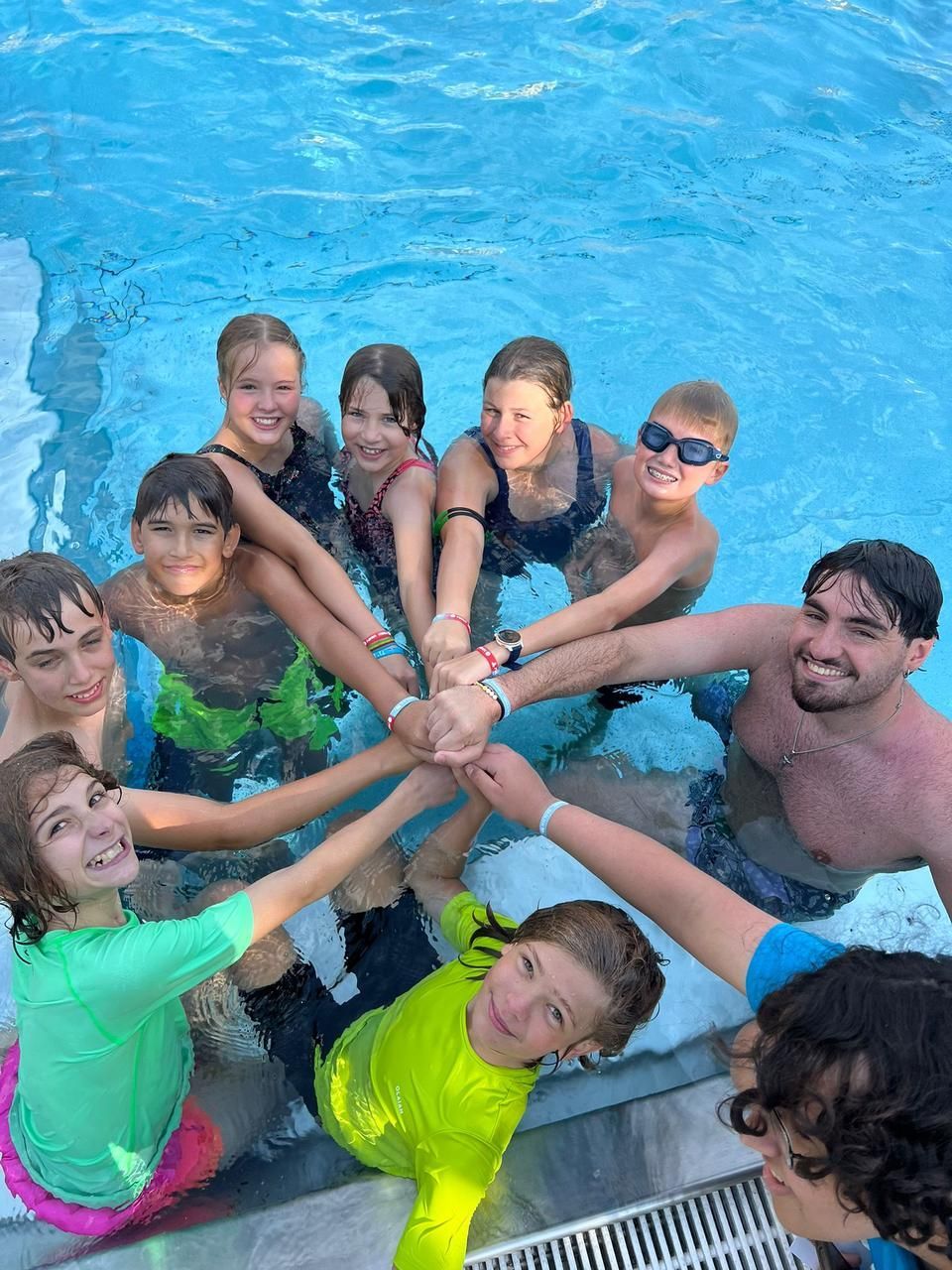 Eine Gruppe von Menschen in einem Schwimmbecken, die Hände im Kreis gefaltet, lächelnd, nahe dem Beckenrand.