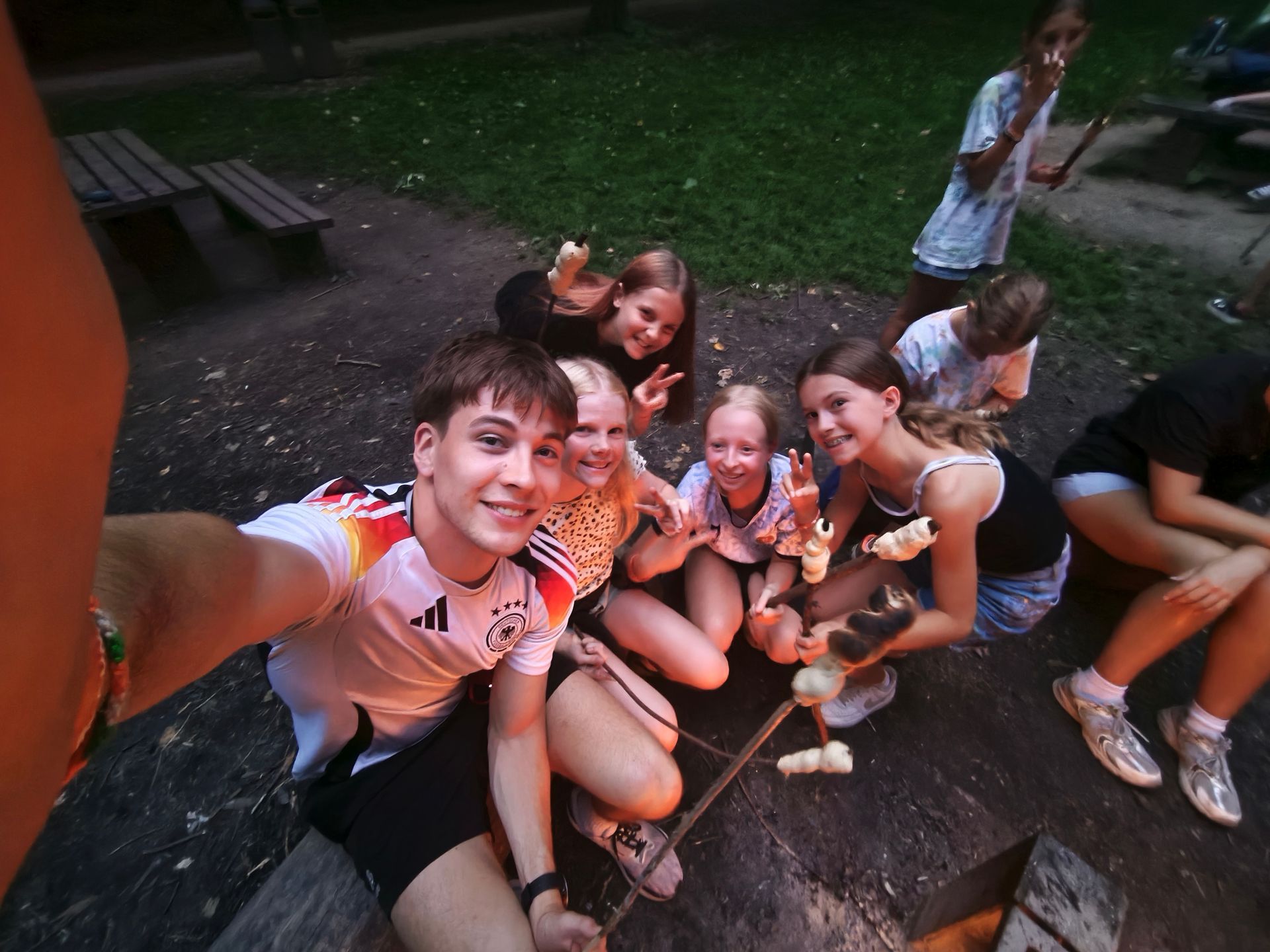 Eine Gruppe von Menschen sitzt um ein kleines Feuer und hält Stöcke mit Essen in den Händen. Sie sitzen im Freien und lächeln.