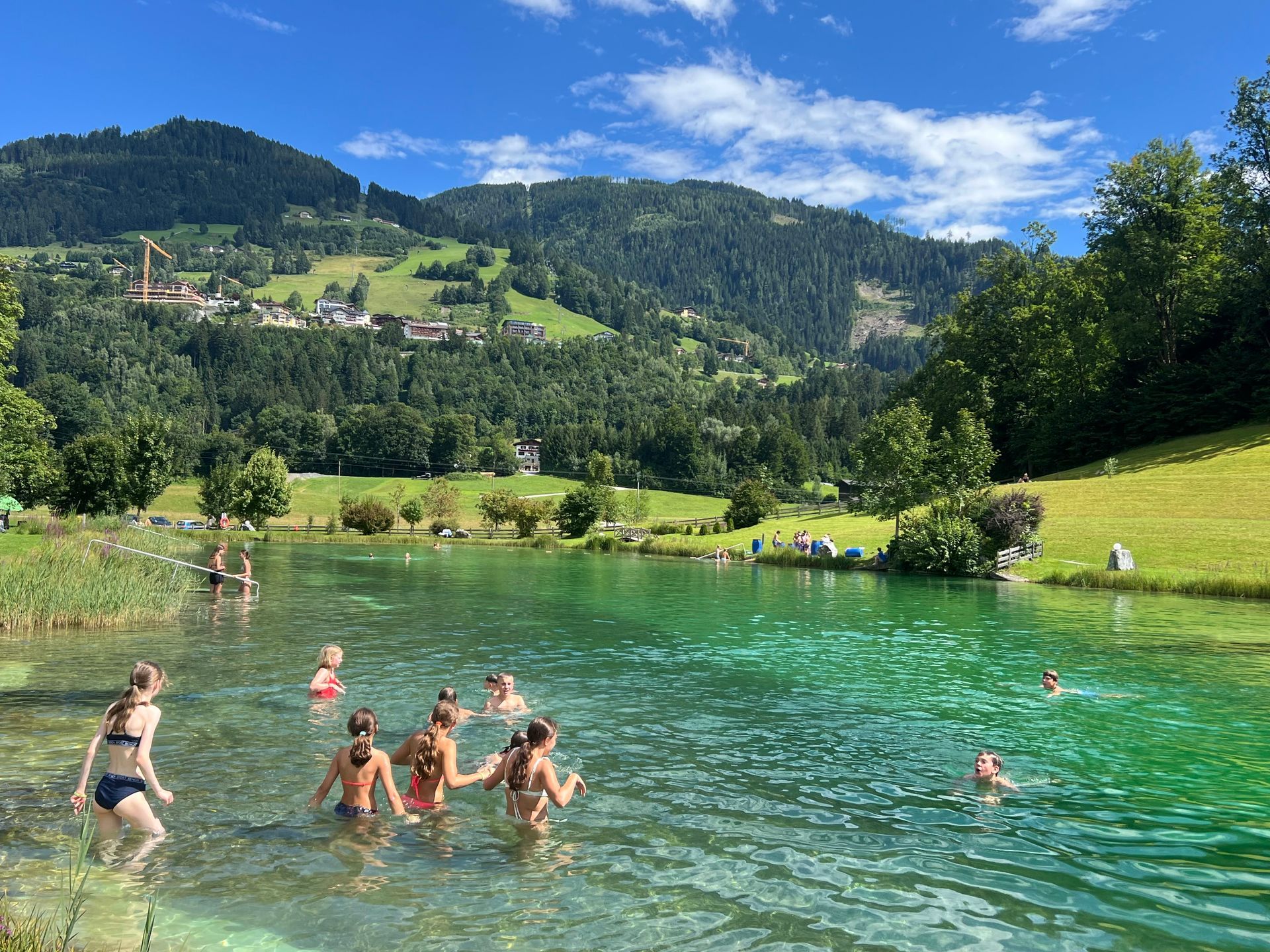 Menschen schwimmen in einem klaren, türkisfarbenen See vor einer Bergkulisse. Sonniger Tag, grünes Gras und Bäume.