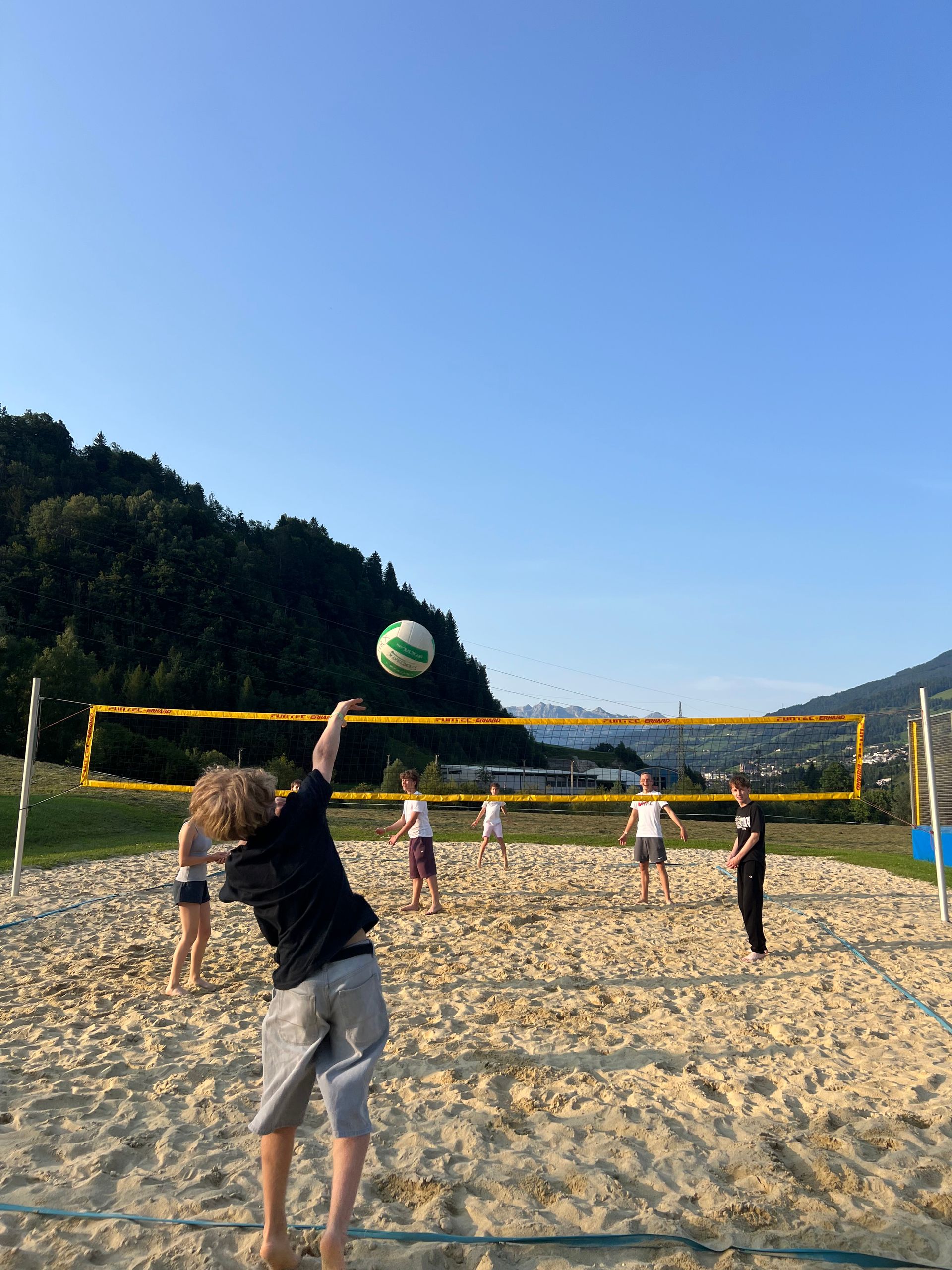 Menschen spielen Volleyball auf einem Sandplatz, der Ball befindet sich in der Luft nahe dem Netz.