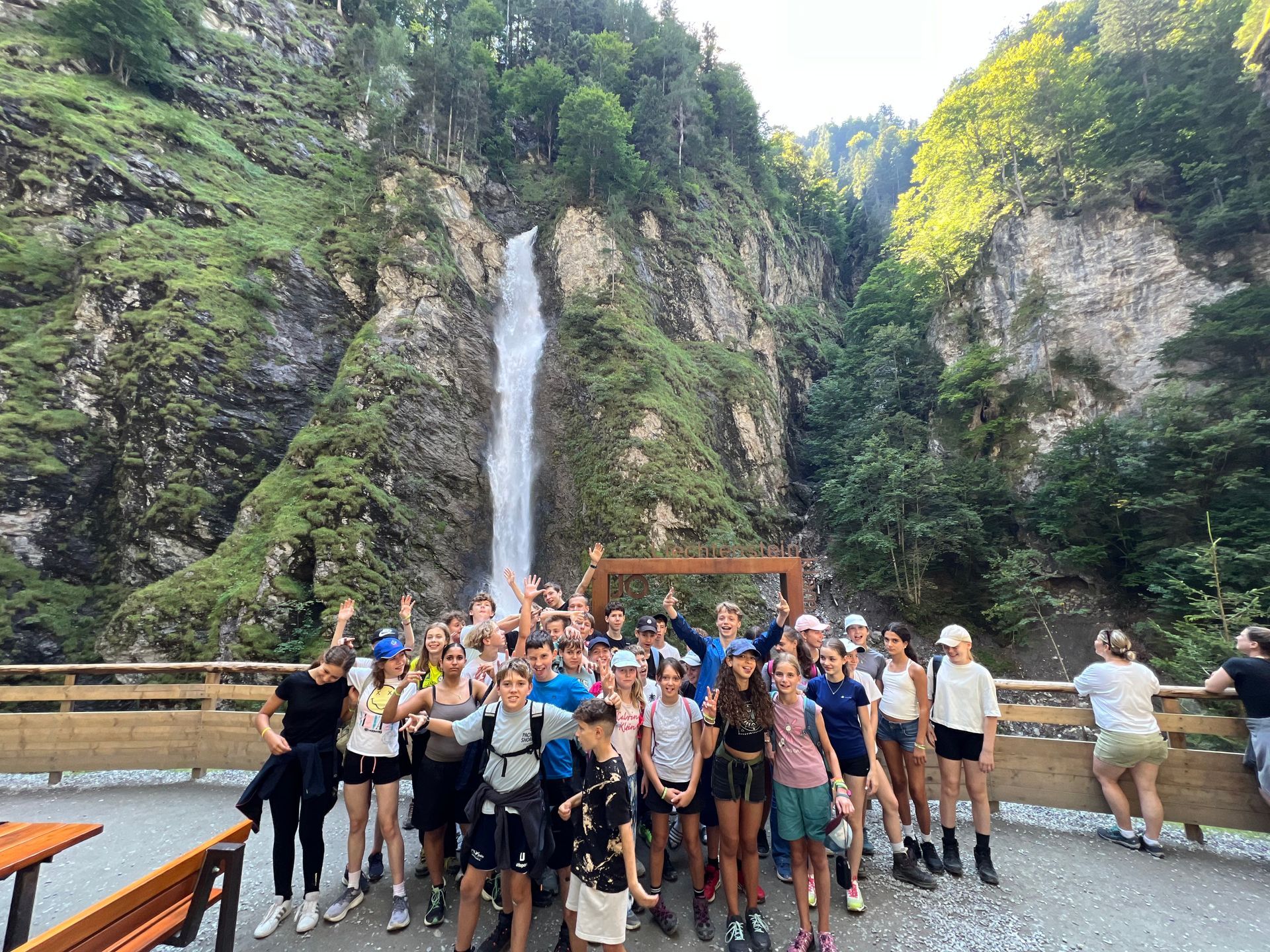 Eine große Menschengruppe posiert vor einem hohen Wasserfall in einem Berggebiet.