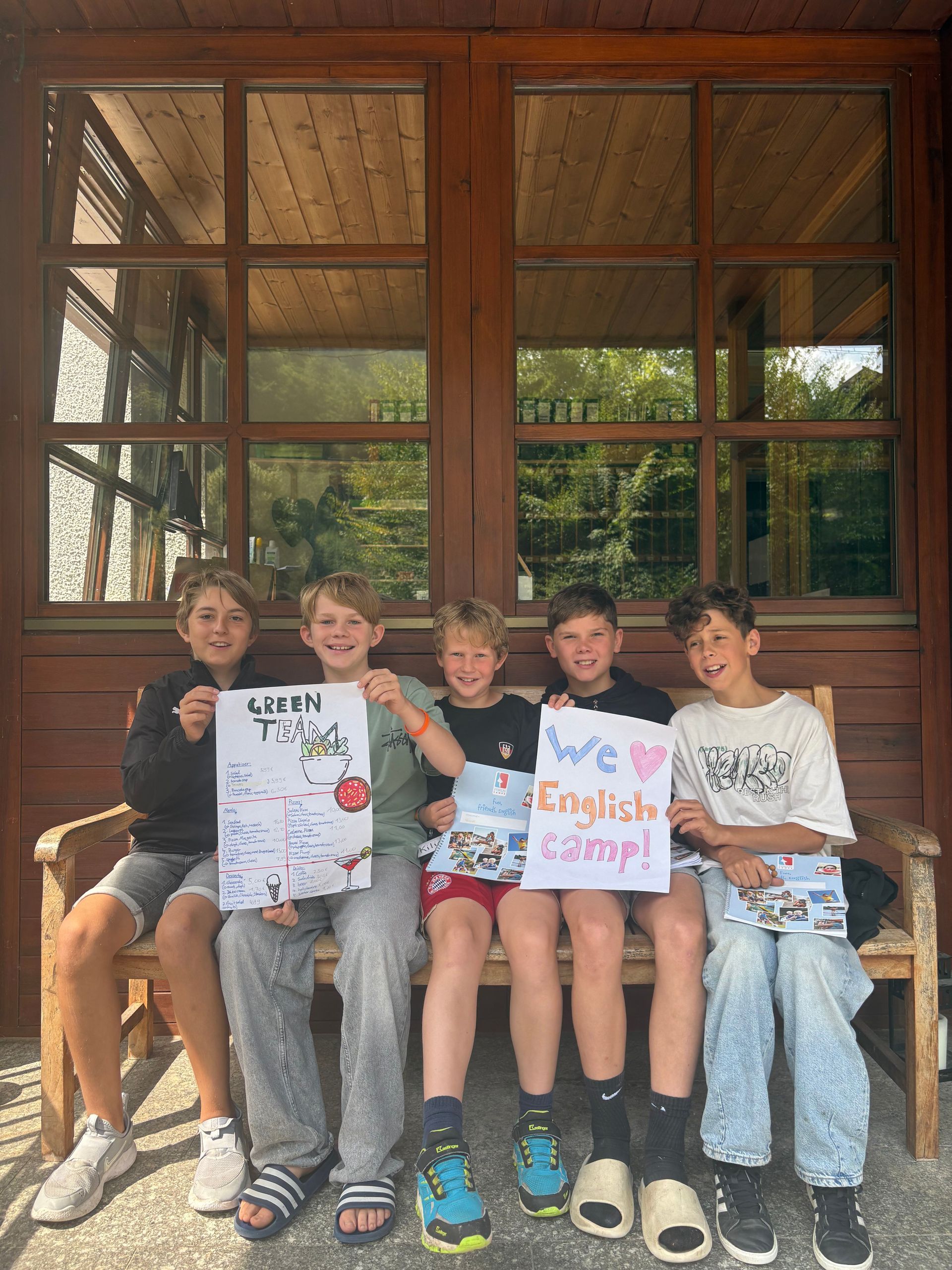 Fünf Jungen sitzen auf einer Holzbank und halten Schilder mit der Aufschrift „Mädchenkram“ und „Wir lieben das Englischcamp!“. Im Hintergrund eine Holzvertäfelung.