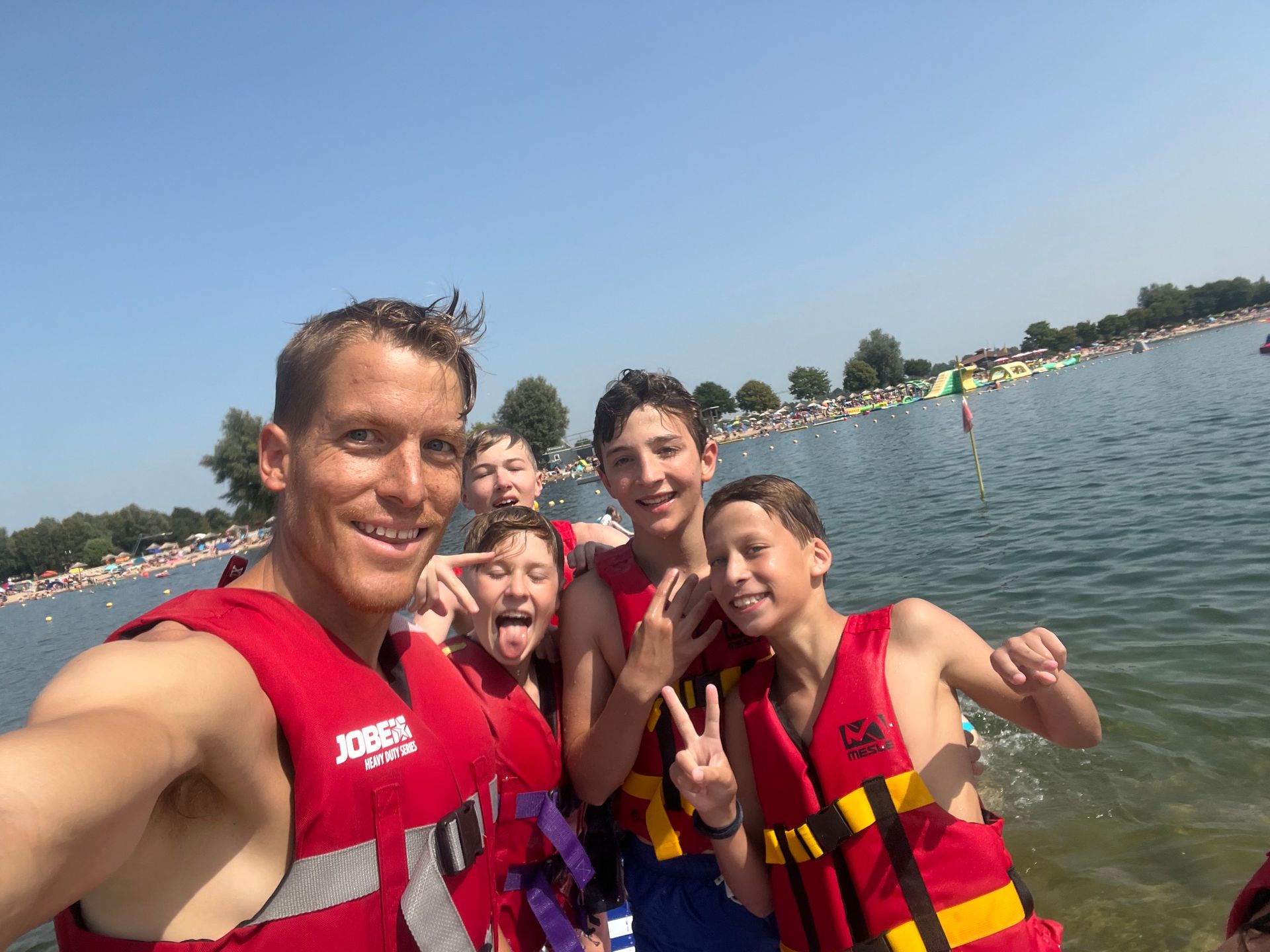 Eine Gruppe von Menschen in roten Schwimmwesten lächelt und gestikuliert auf einem See. Im Hintergrund strahlend blauer Himmel.