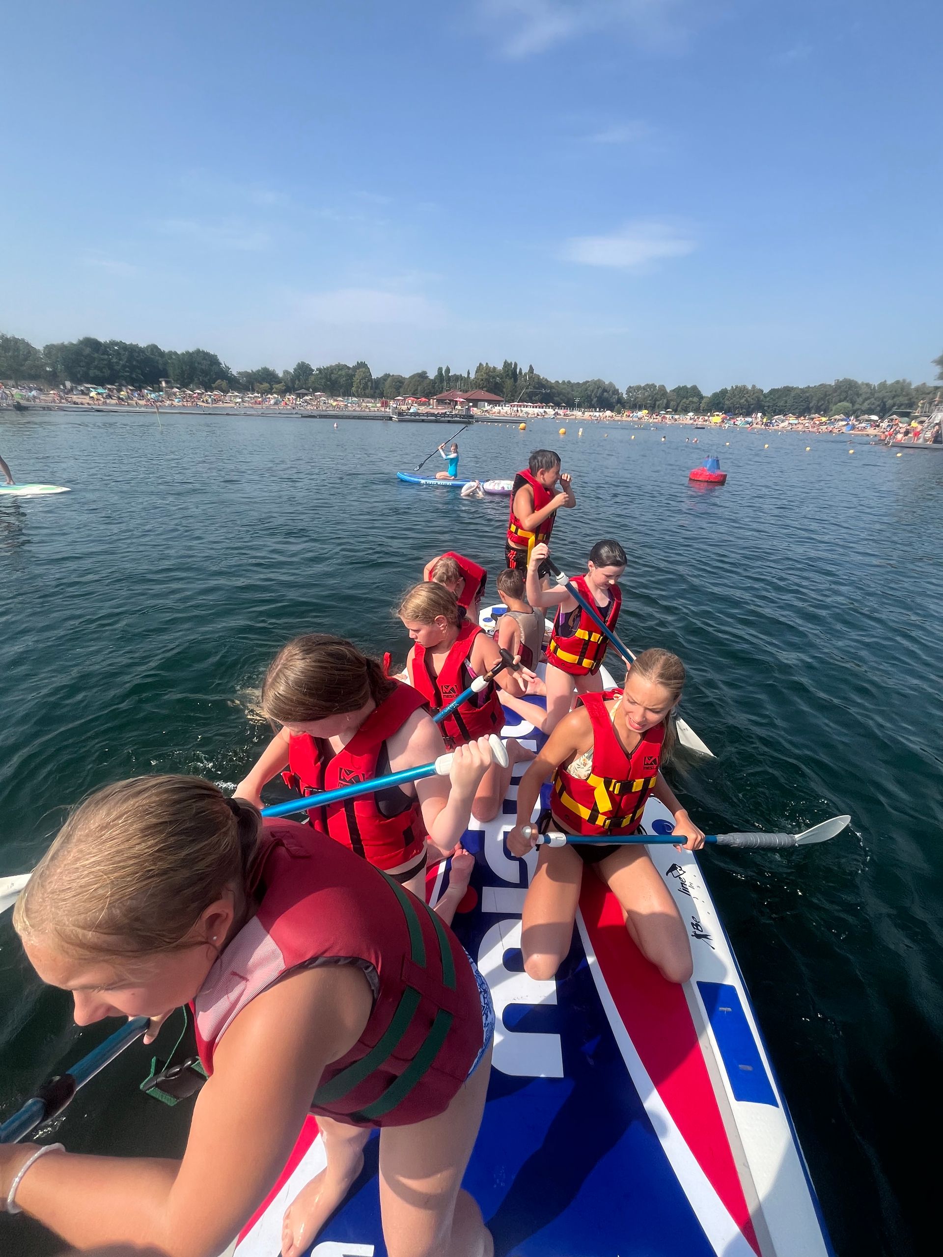 Kinder in Schwimmwesten paddeln auf einem großen Paddleboard auf einem See.