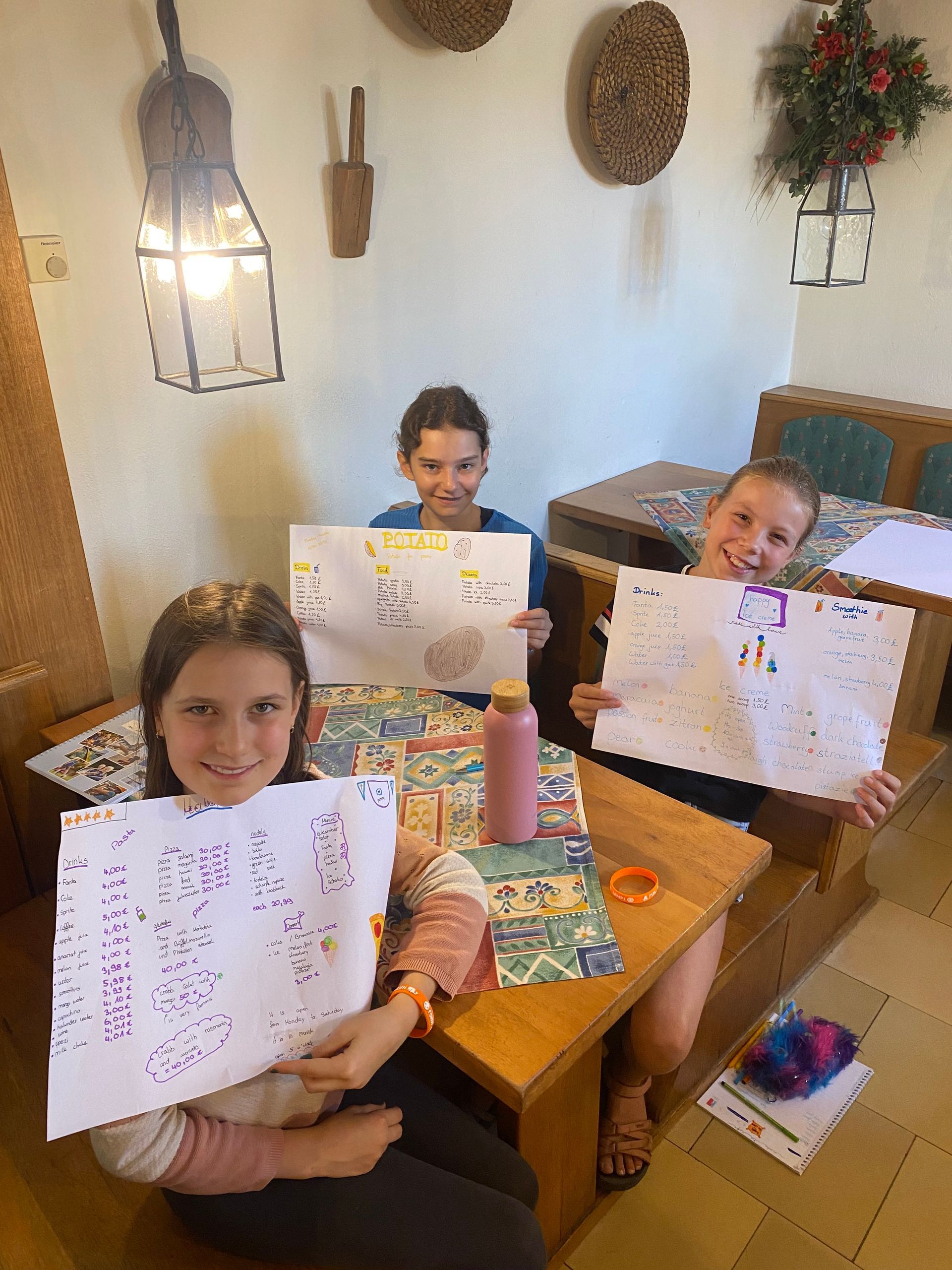 Drei Kinder sitzen lächelnd an einem Tisch und halten Zeichnungen hoch. Helles Zimmer mit Holztisch und Wanddekoration.