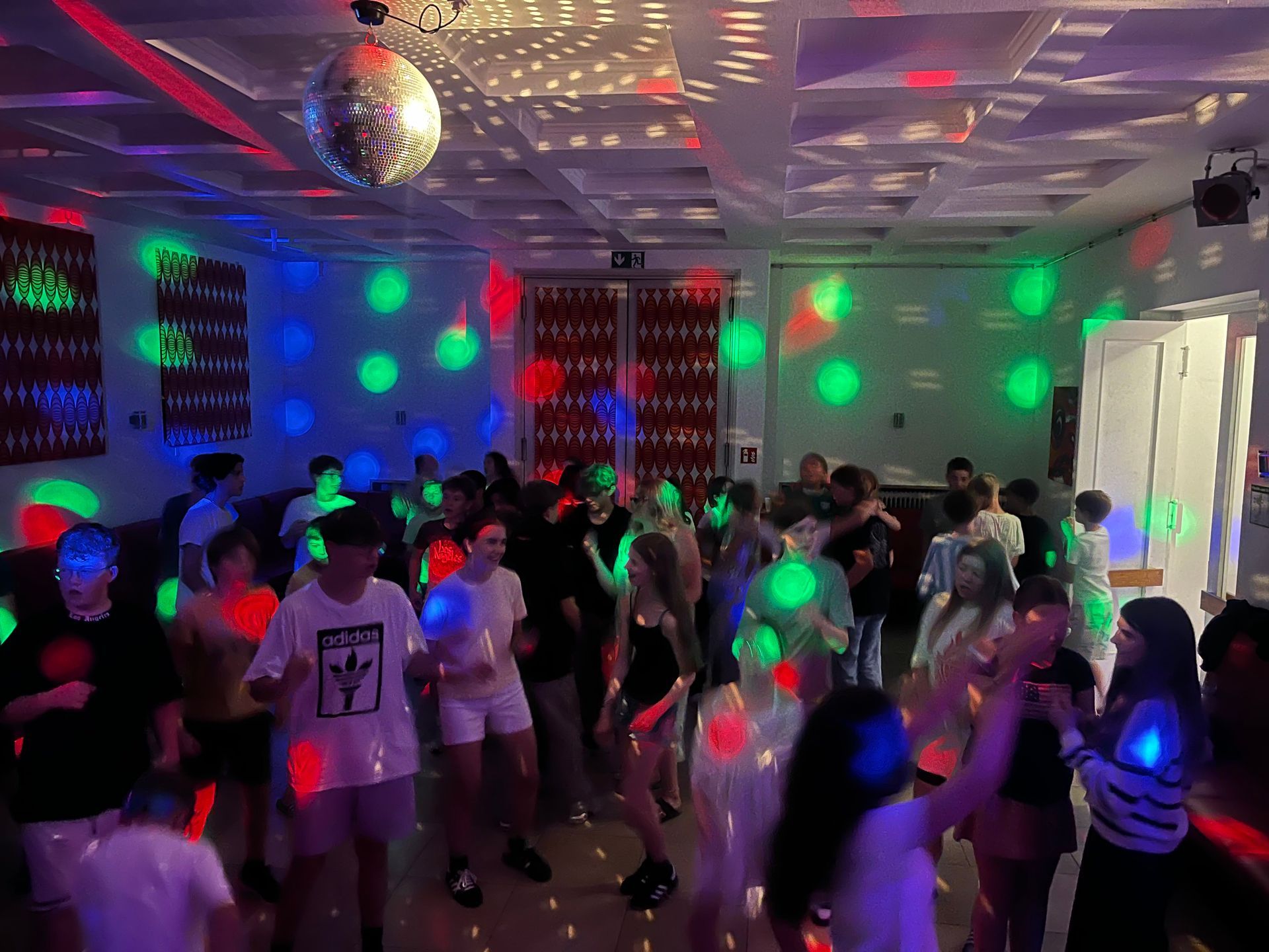 Menschen tanzen in einem Raum, der von einer Discokugel und bunten Lichtern erhellt wird.