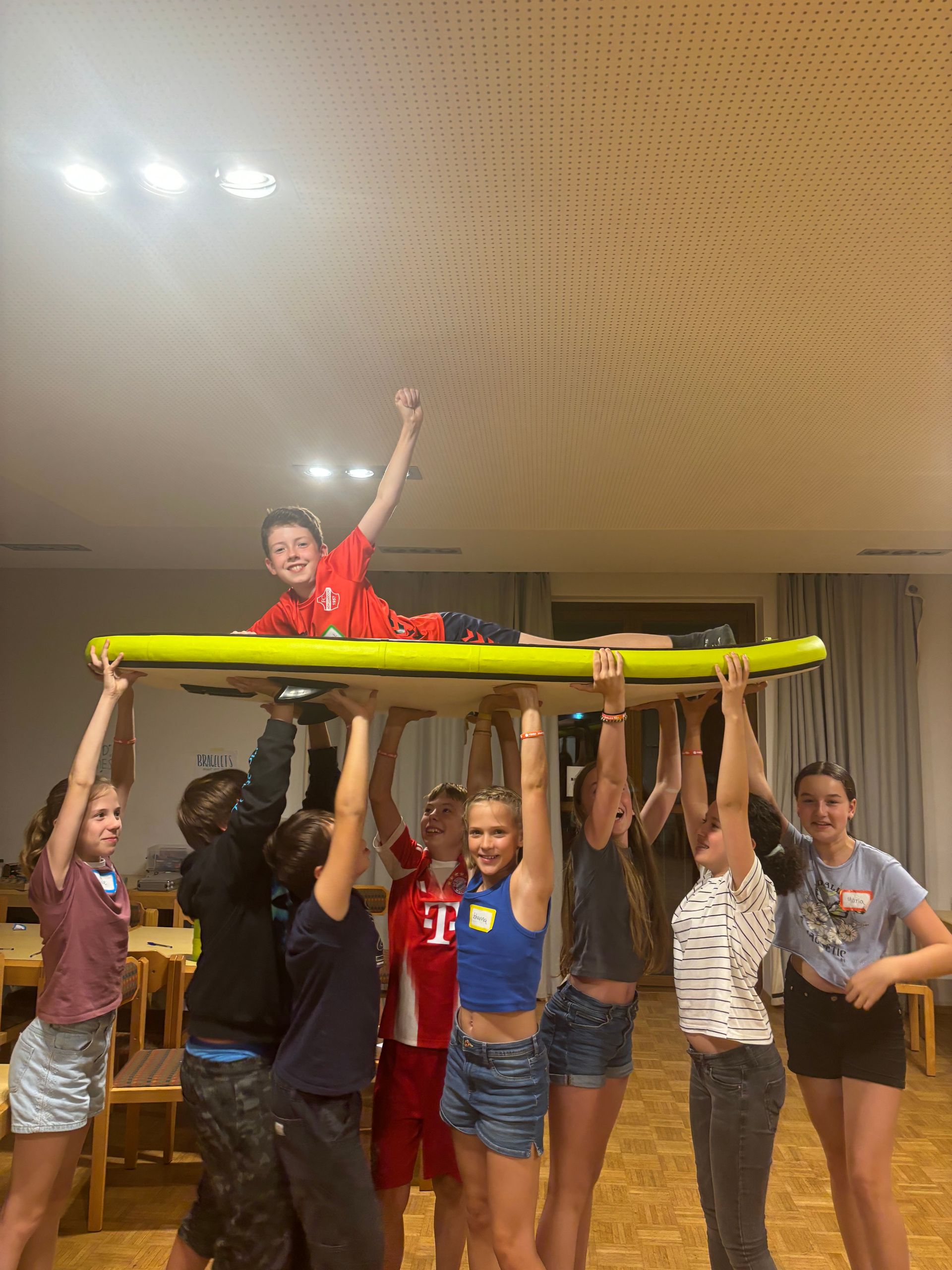 Eine Gruppe Kinder stützt einen Jungen auf einem Holzbrett. Der Junge hebt den Arm. Innenbereich, Holzboden.