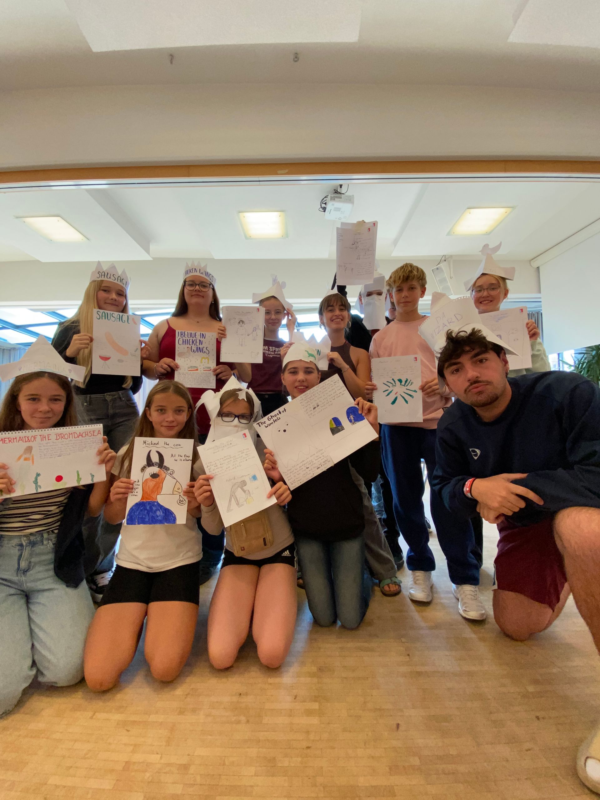 Eine Gruppe Kinder mit Papierkronen hält Zeichnungen in den Händen und lächelt. Drinnen knien einige.