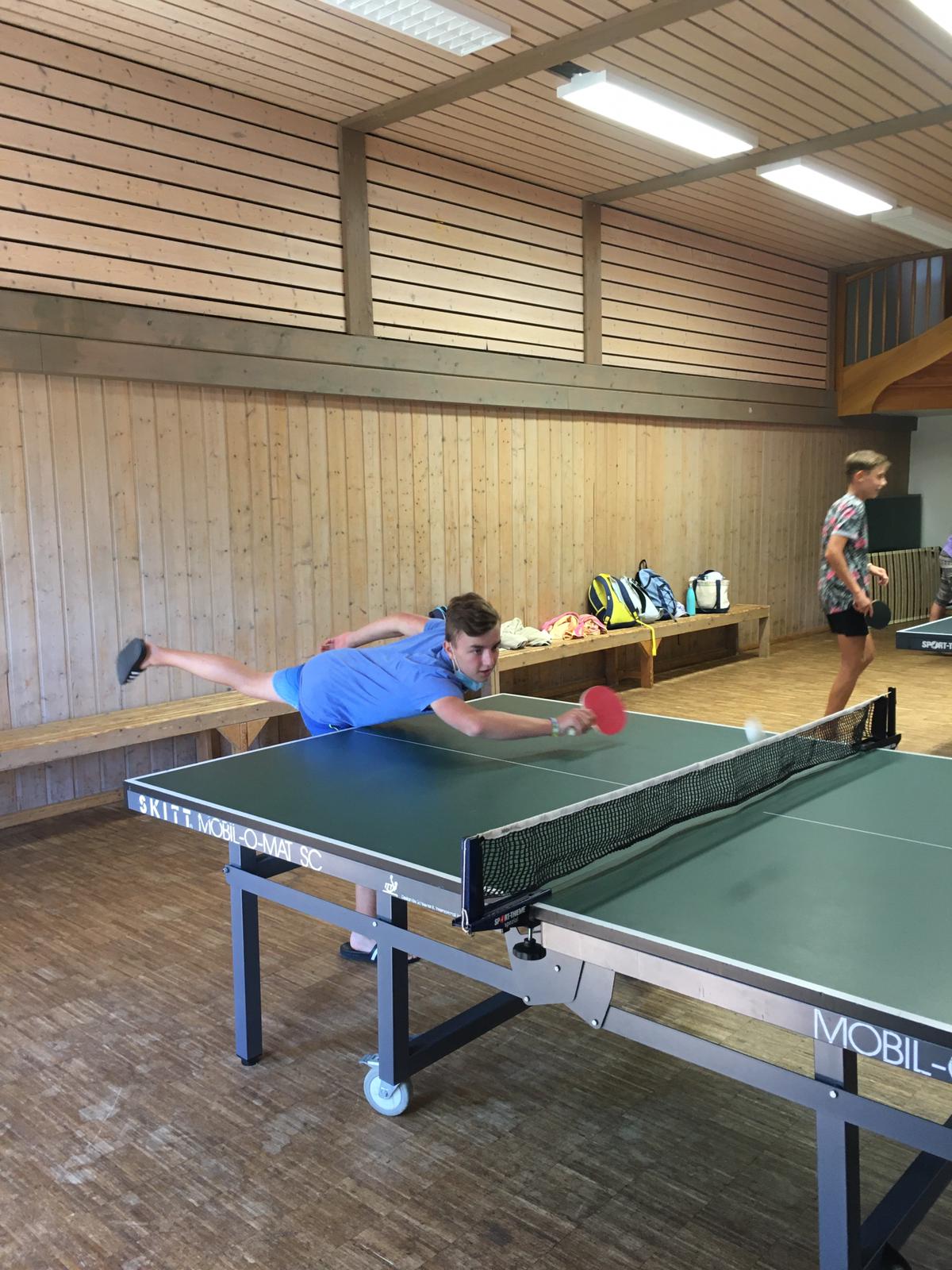 Eine Person hechtet nach einem Tischtennisball. Eine andere Person beobachtet das Spiel in einem holzgetäfelten Raum.