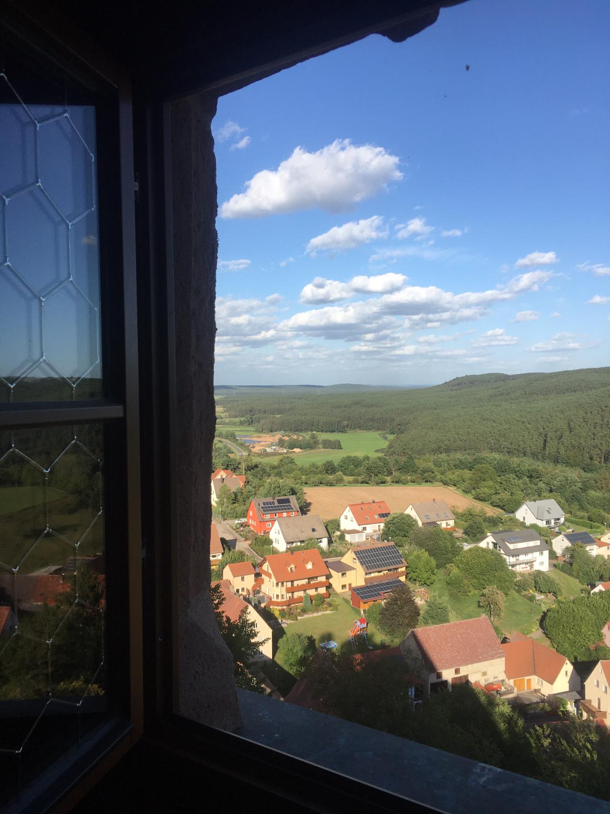 Blick aus einem Schlossfenster auf ein kleines Dorf mit roten Ziegeldächern und grünen Bäumen unter blauem Himmel.