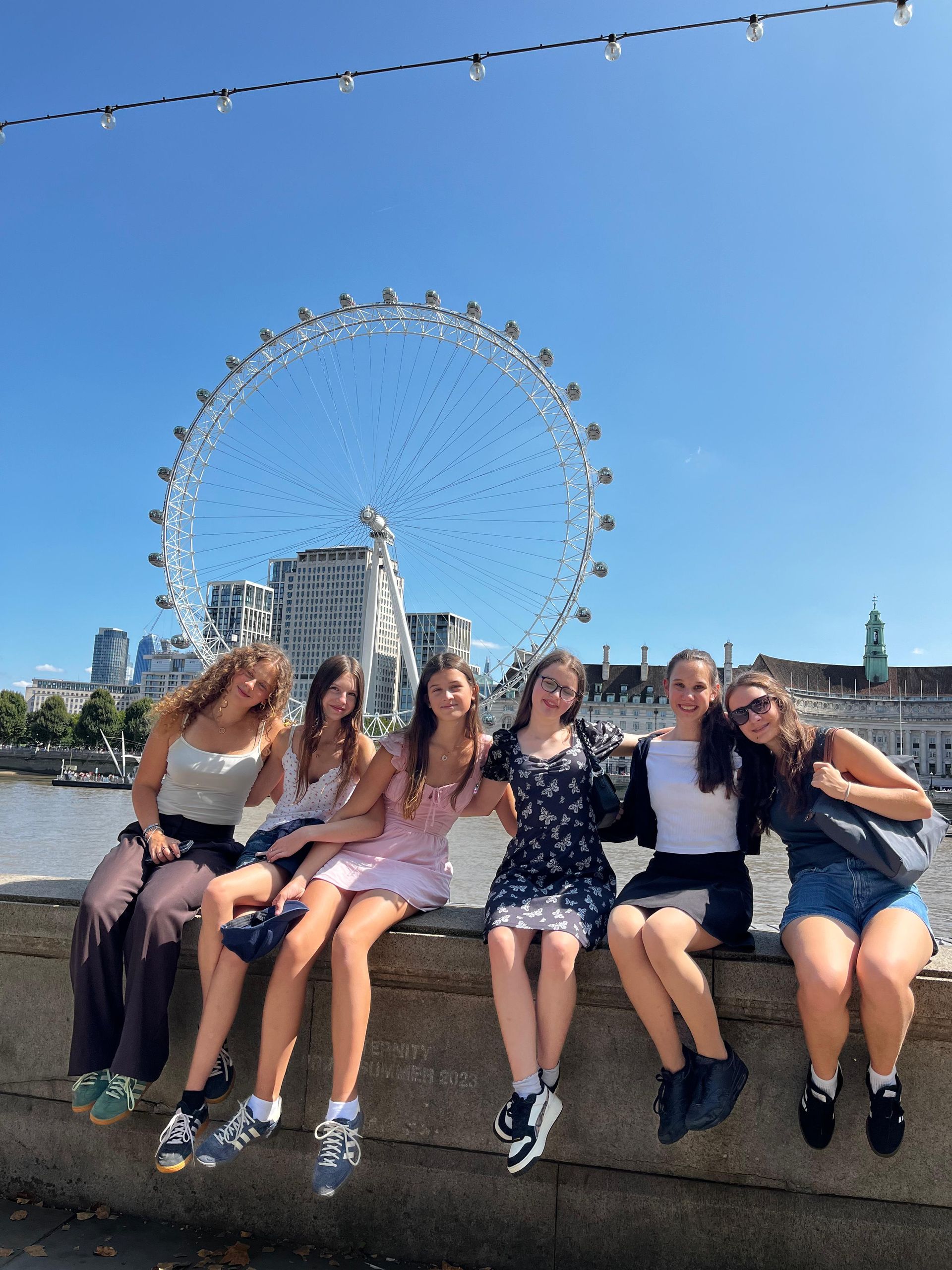 Sechs Personen sitzen auf einer Mauer vor dem London Eye. Sonniger Tag; klarer Himmel.