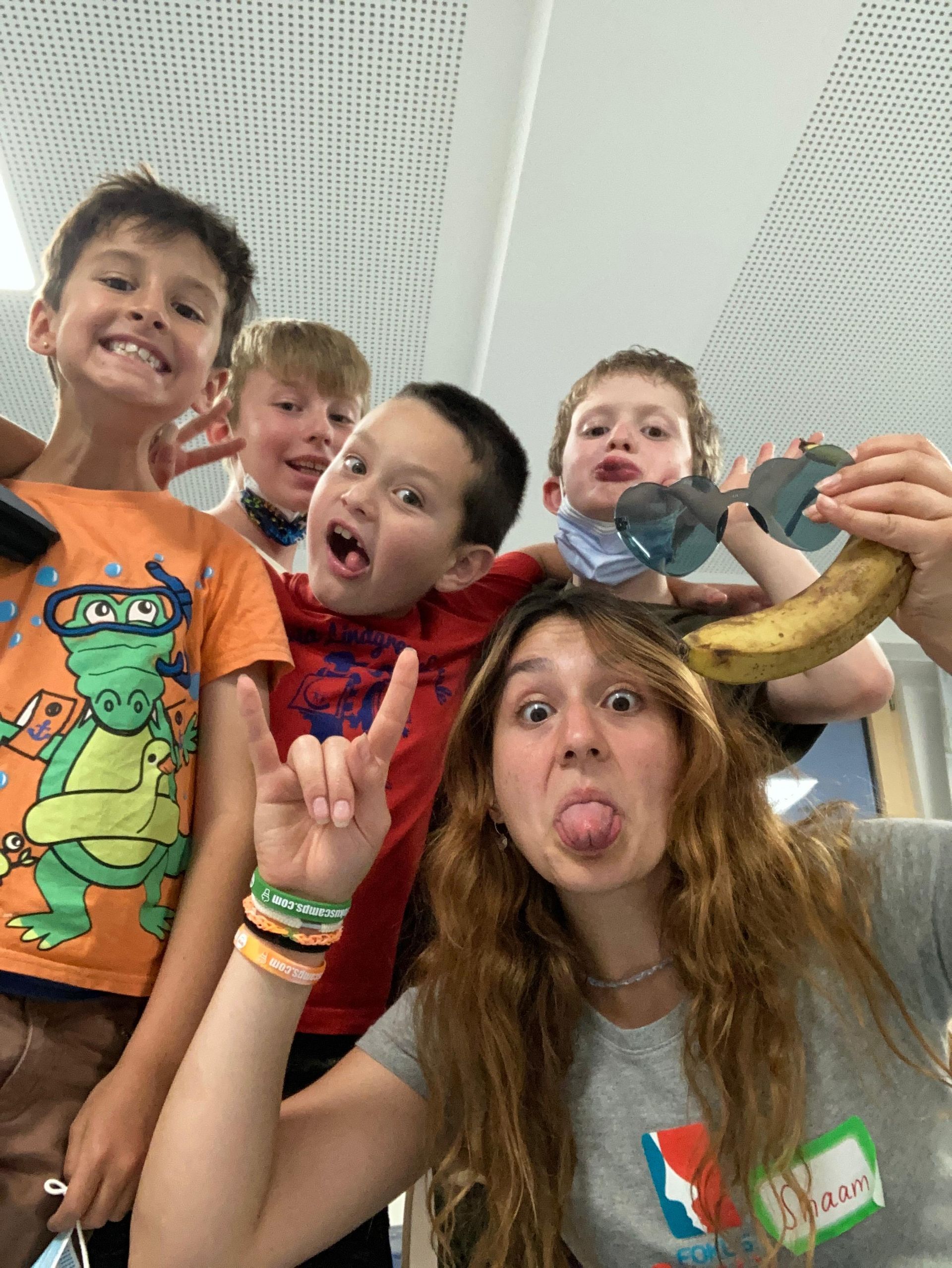 Eine Gruppe Kinder und eine Frau schneiden Grimassen und halten eine Banane in der Hand; sie tragen eine Sonnenbrille. (Indoor)