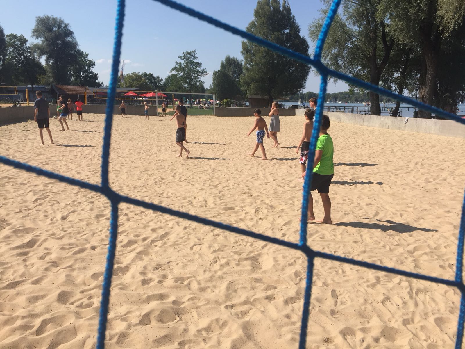 Menschen spielen Beachvolleyball auf einem sandigen Platz, beobachtet durch ein blaues Netz an einem sonnigen Tag.