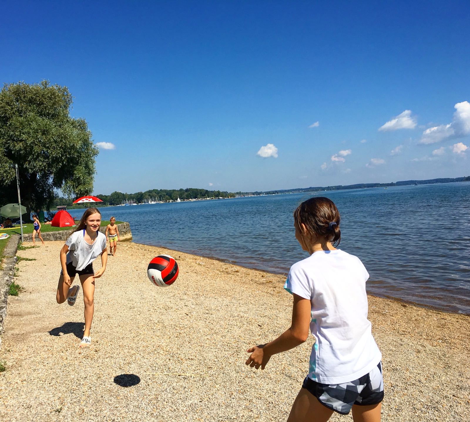Zwei Mädchen spielen an einem sonnigen Tag an einem Strand an einem See mit einem rot-schwarzen Ball.
