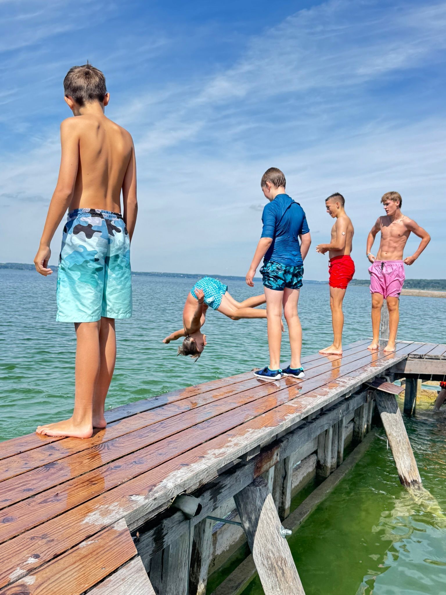 Fünf Personen auf einem Holzsteg; eine macht einen Rückwärtssalto ins Wasser. Blaues Wasser, sonniger Himmel.