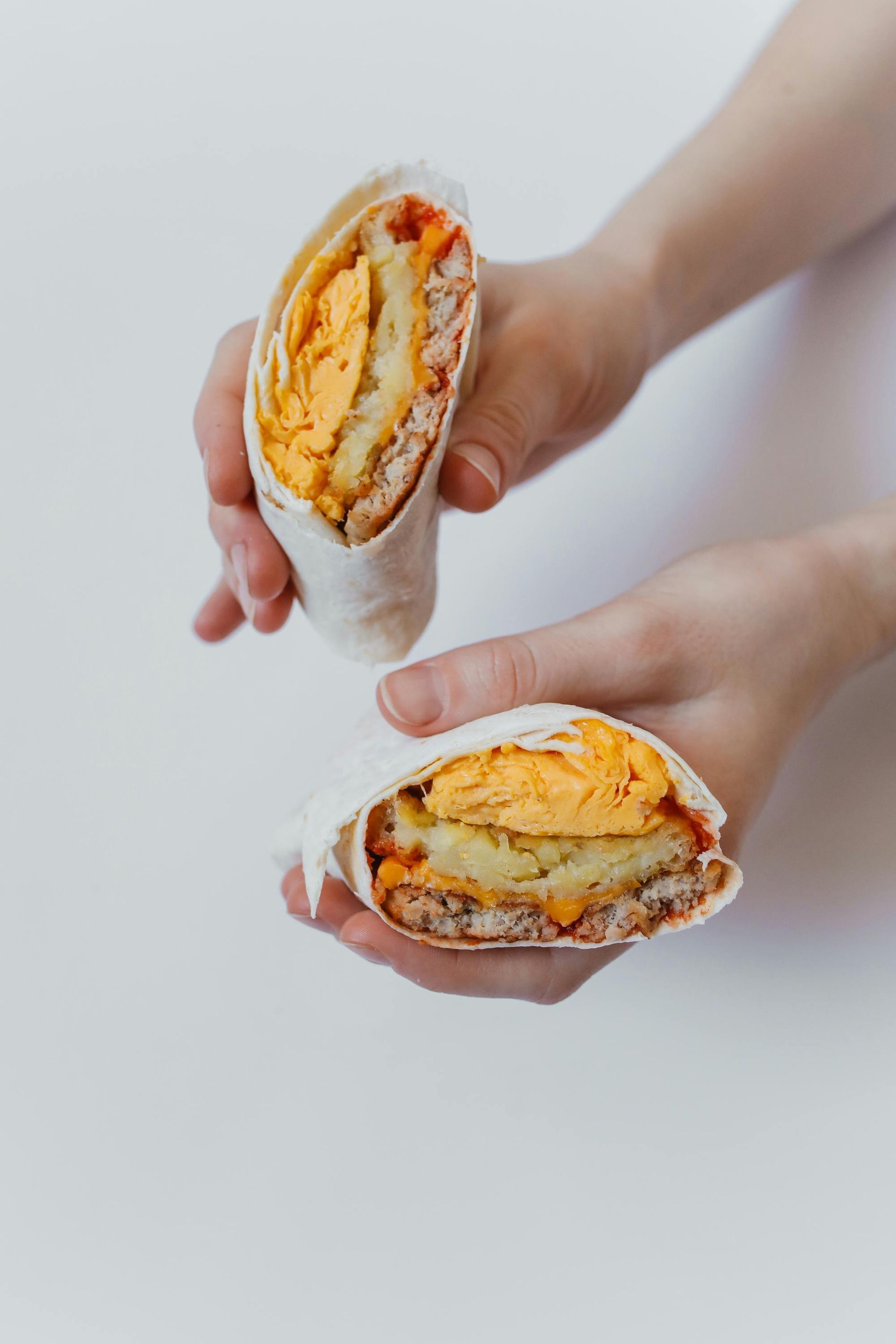 Zwei Hände halten Hälften eines eingewickelten Frühstücks-Burritos, aus dem Eier, Kartoffeln und rote Soße hervortreten.