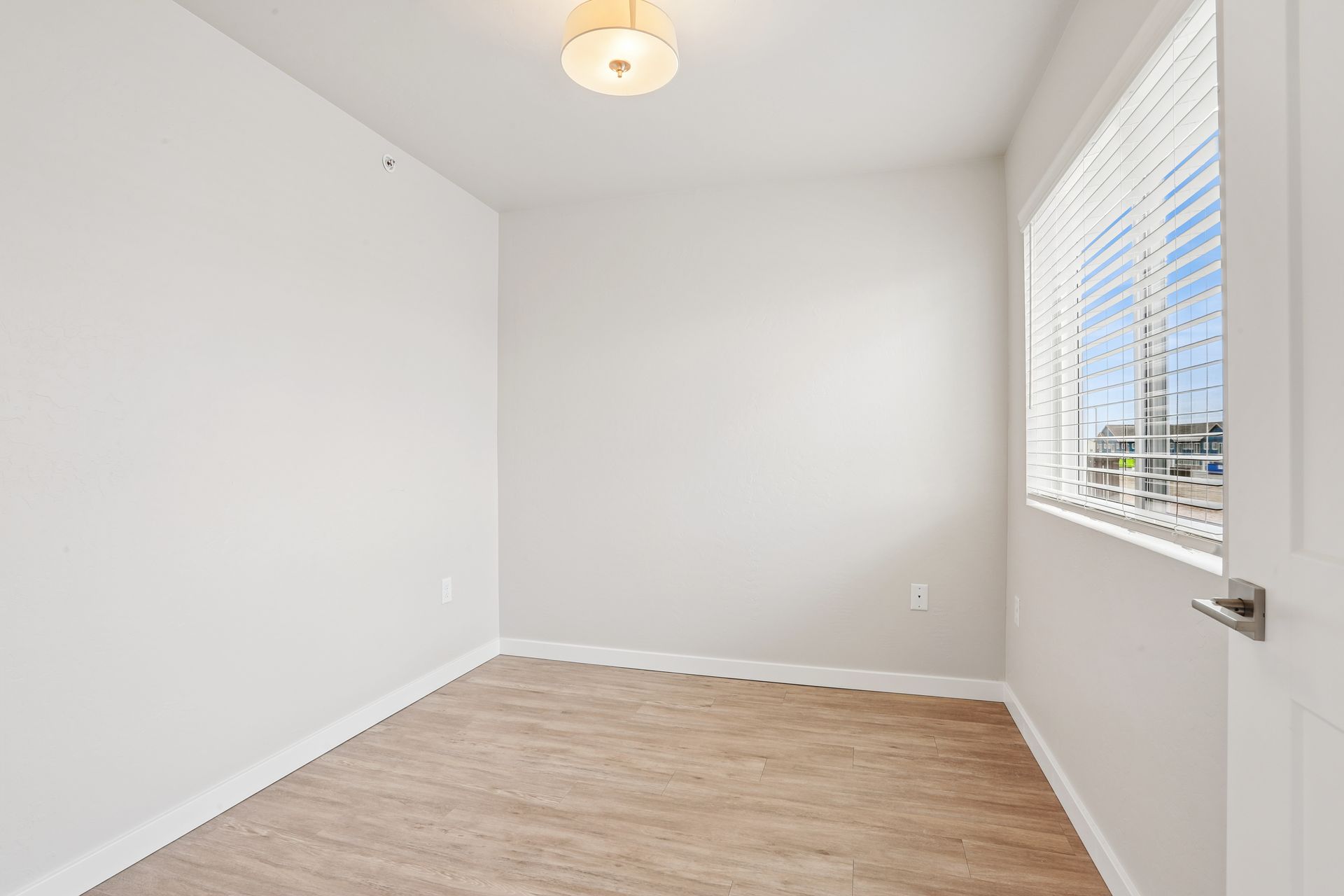 Empty white bedroom with wood-look floor, ceiling light, and a window with blinds.