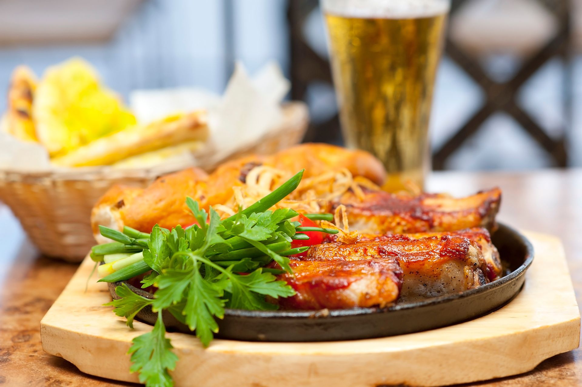 A close-up shot of a sizzling hot cast-iron skillet filled with grilled, glazed meat, topped with fried onion straws and fresh green herbs like parsley and chives. A tall, cold glass of beer sits in the blurred background.