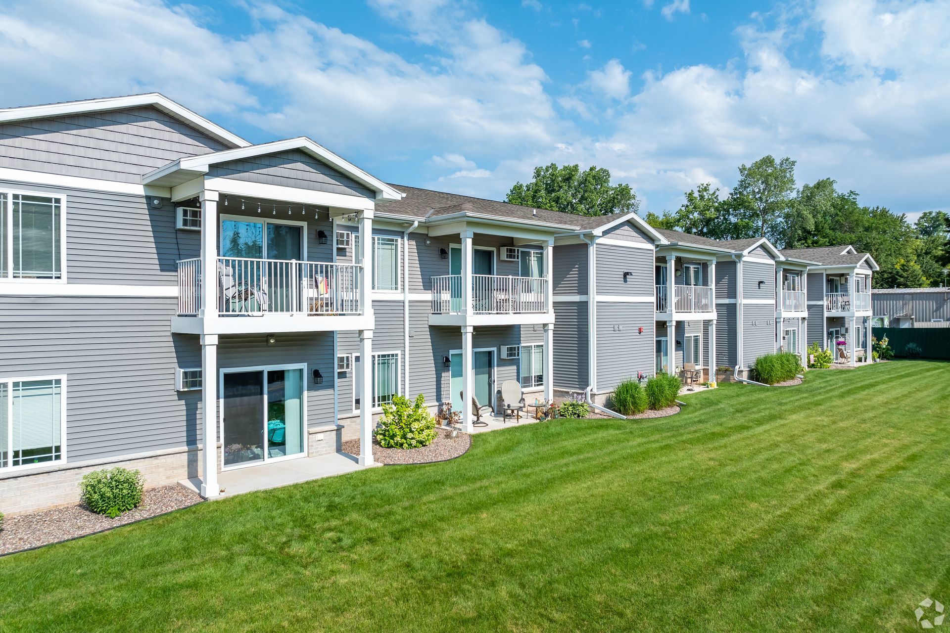 Row of gray apartment buildings with balconies beside a green lawn under a blue sky