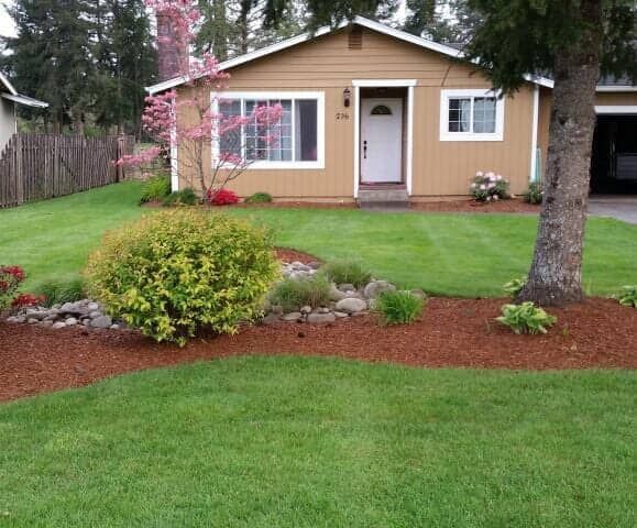 Small tan house with white trim, front lawn, flower beds, and trees in a suburban yard.