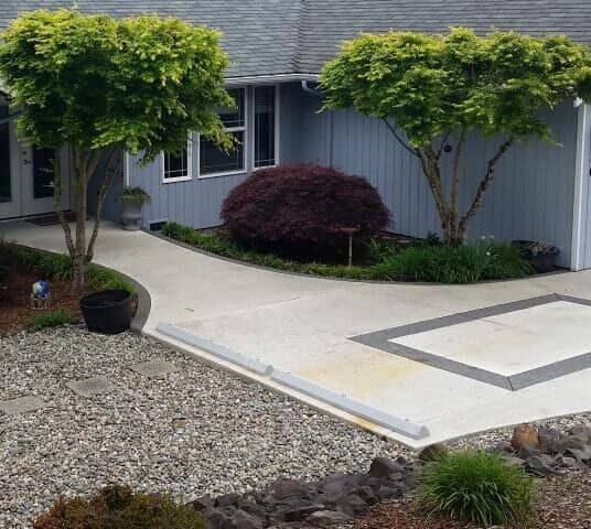 Front yard with a curved concrete walkway, gravel, and neatly trimmed trees beside a gray house