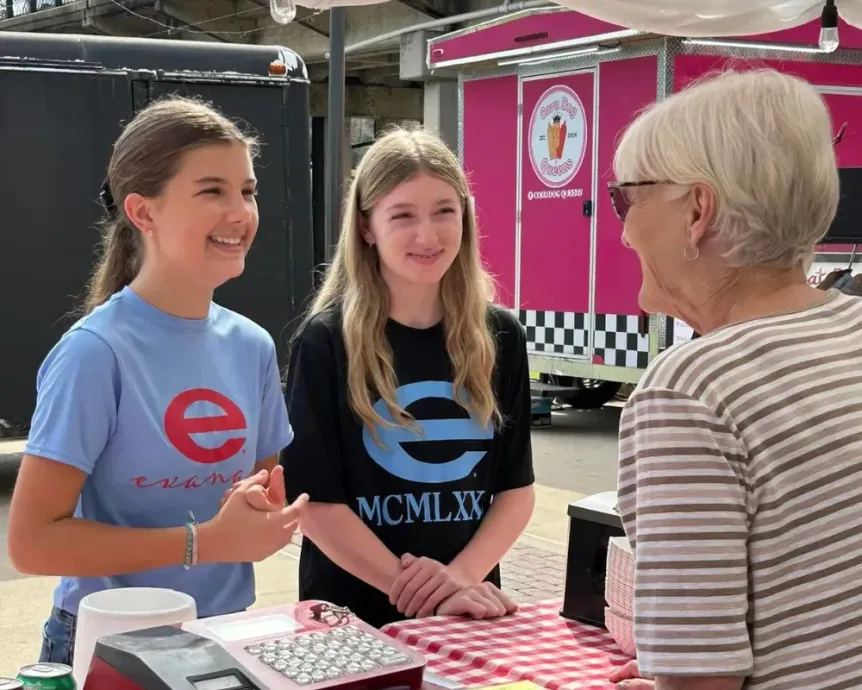 Two volunteers at a checkered table talking with an older woman beside a pink food truck