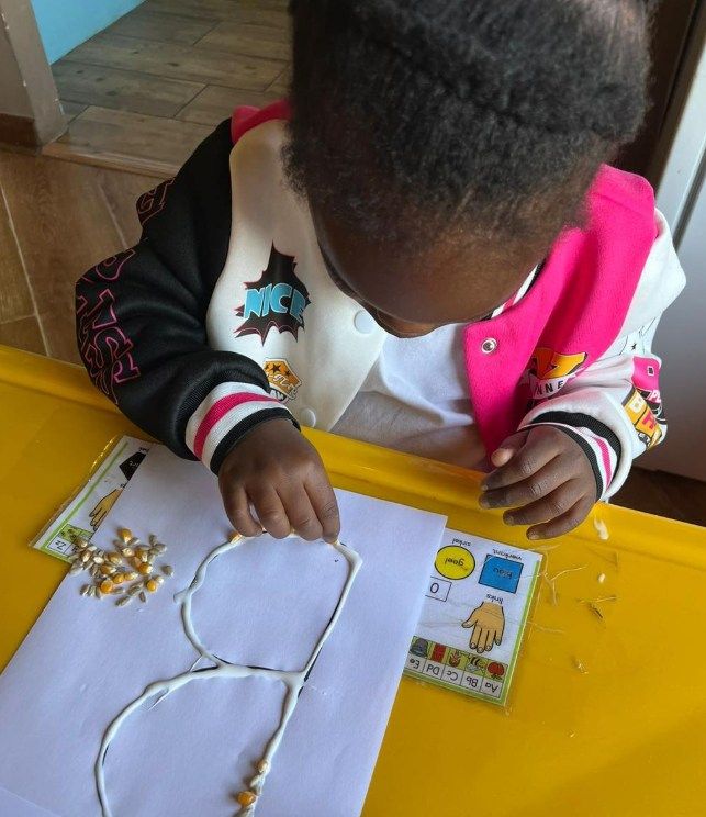 Child arranging corn kernels on a paper outline, at a yellow table.