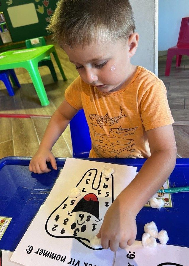 Boy placing cotton balls on a number matching worksheet at a table. He is in a classroom.