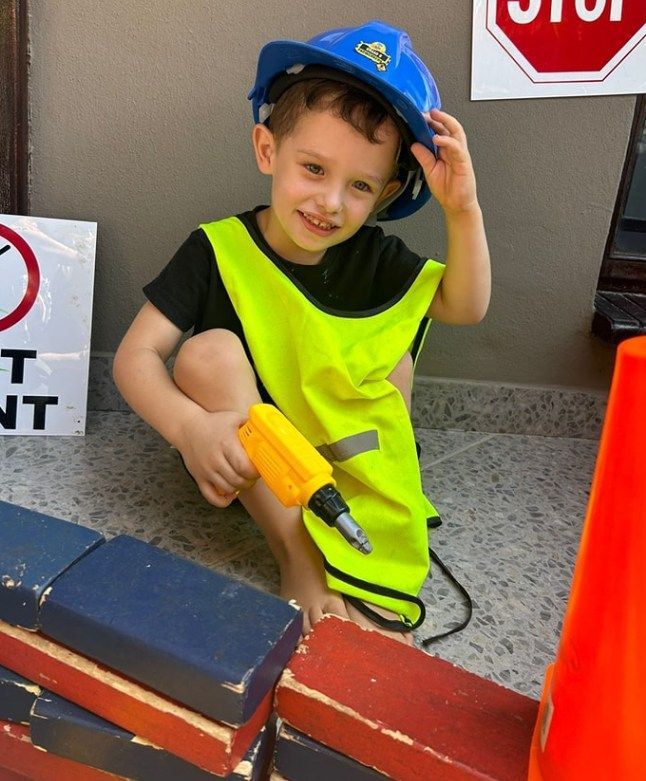 Child dressed as a construction worker, smiling, wearing a hard hat, reflective vest, and holding a toy drill.