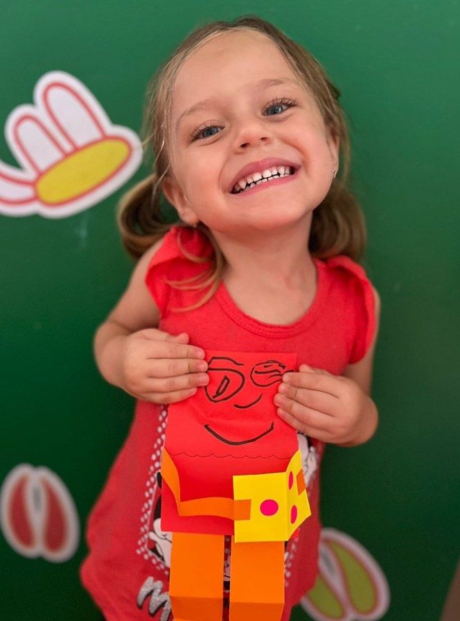 Smiling child in red shirt, holding up a paper figure. Green backdrop with cartoon images.