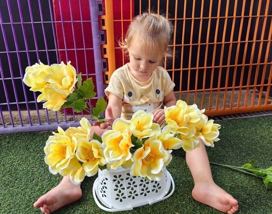 Young child sitting with a basket of yellow flowers, colorful fence background.
