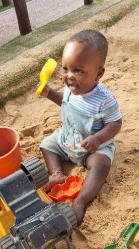 Child playing in sand with toys; smiling, holding a yellow shovel.