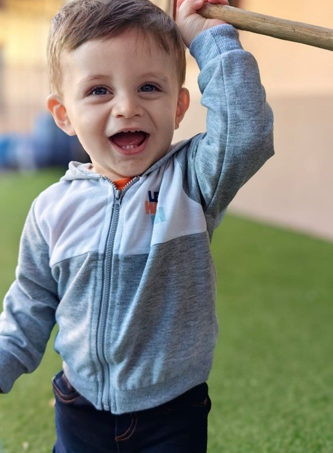Smiling toddler holding a stick, wearing a gray and white hooded jacket, on green grass.