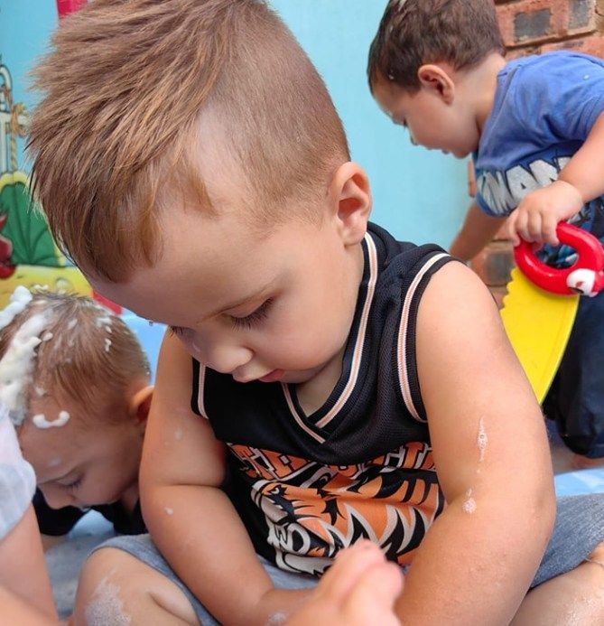 Boy in a tank top sits looking down. Two other children play nearby.