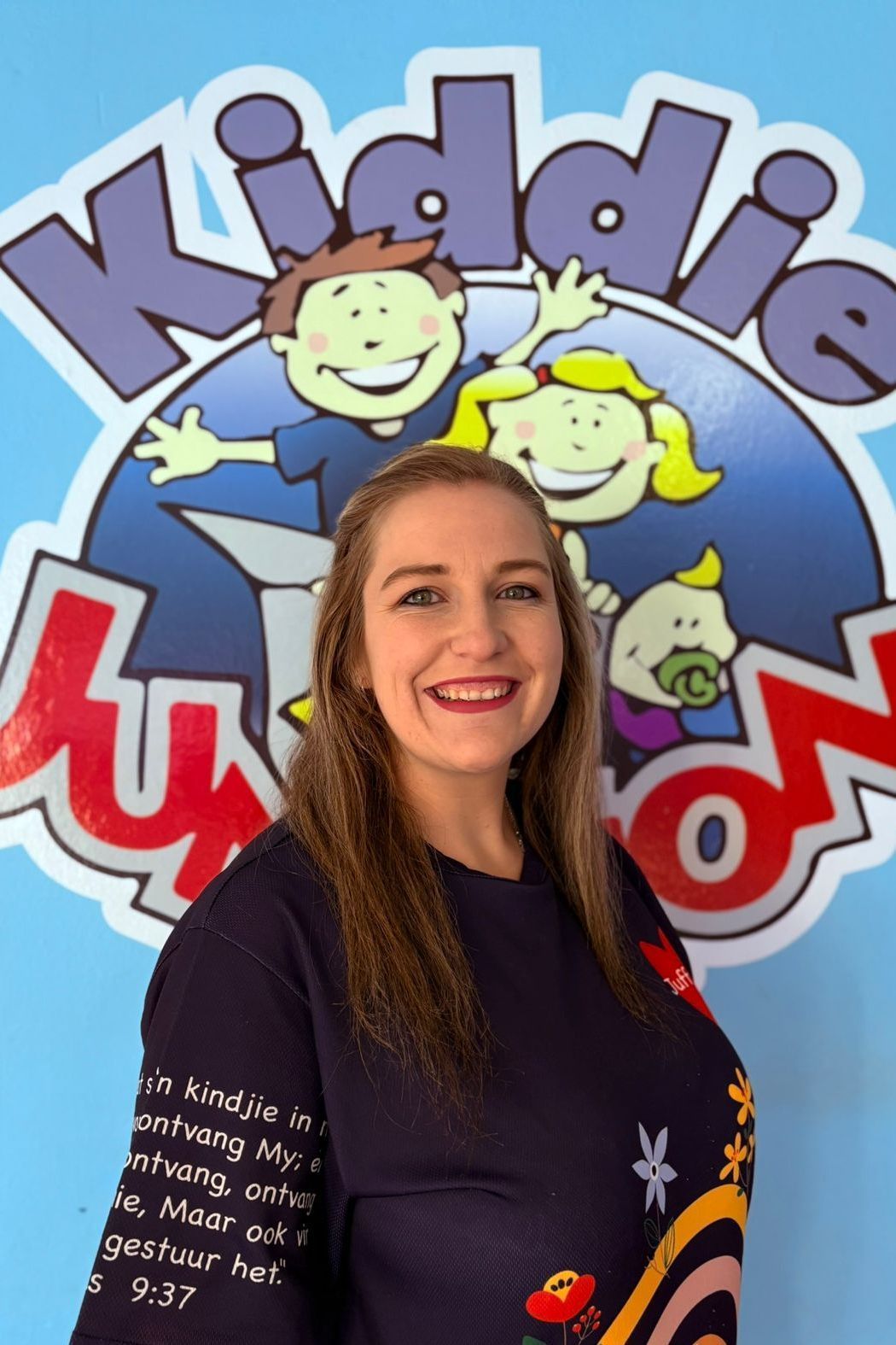Woman smiling in front of a blue Kiddie Nation logo. She wears a navy sweater with colorful floral design.