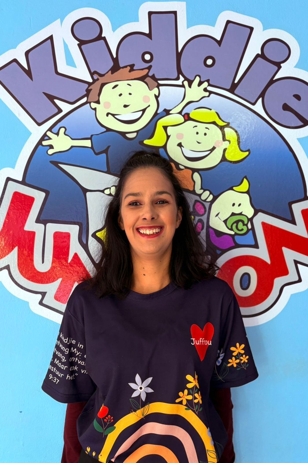 Woman in a decorated blue shirt smiles in front of a blue Kiddie Luxon logo.